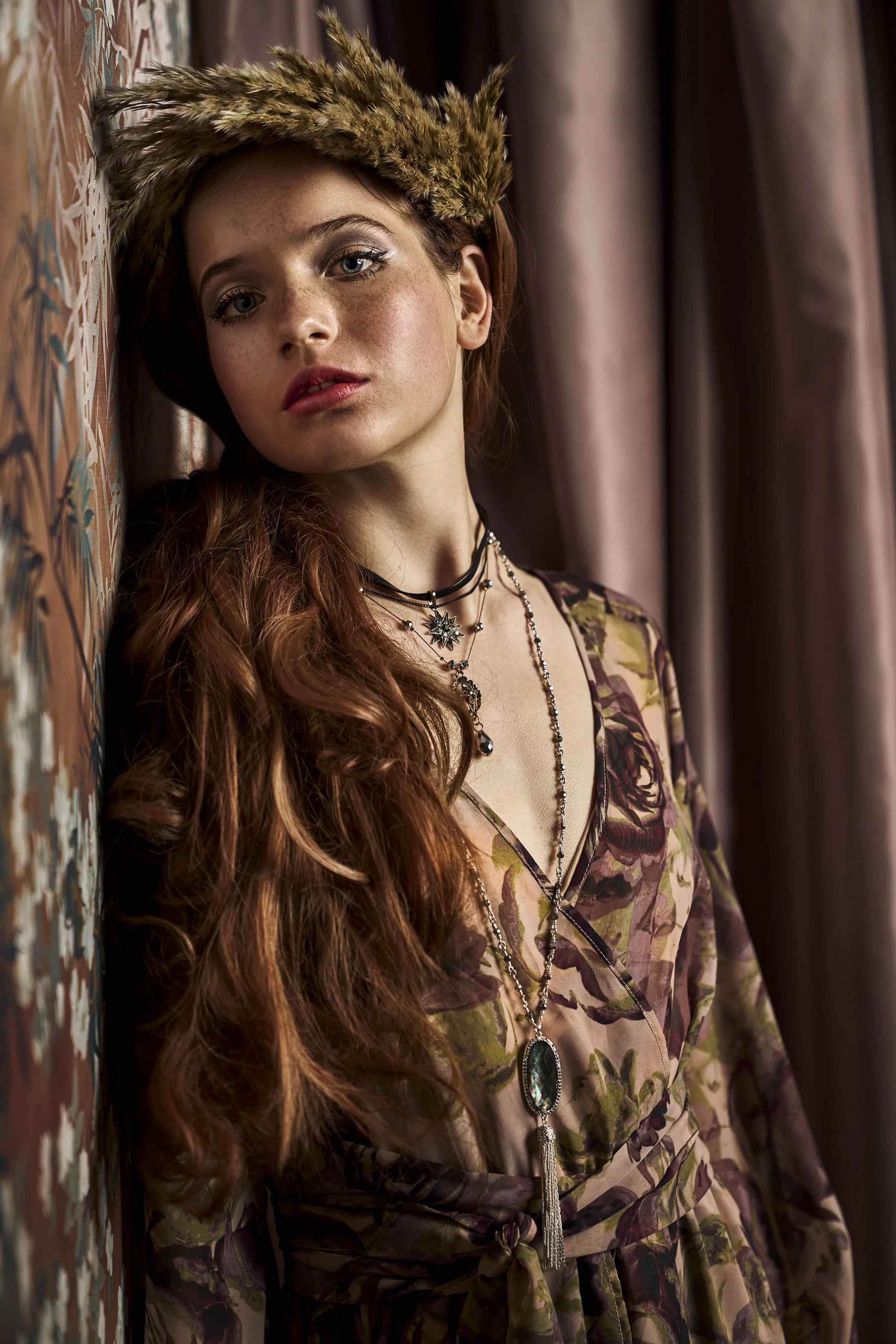 A woman with long red hair and freckles leaning against a decorated wall, wearing a floral dress and layered necklaces with a large gemstone pendant.