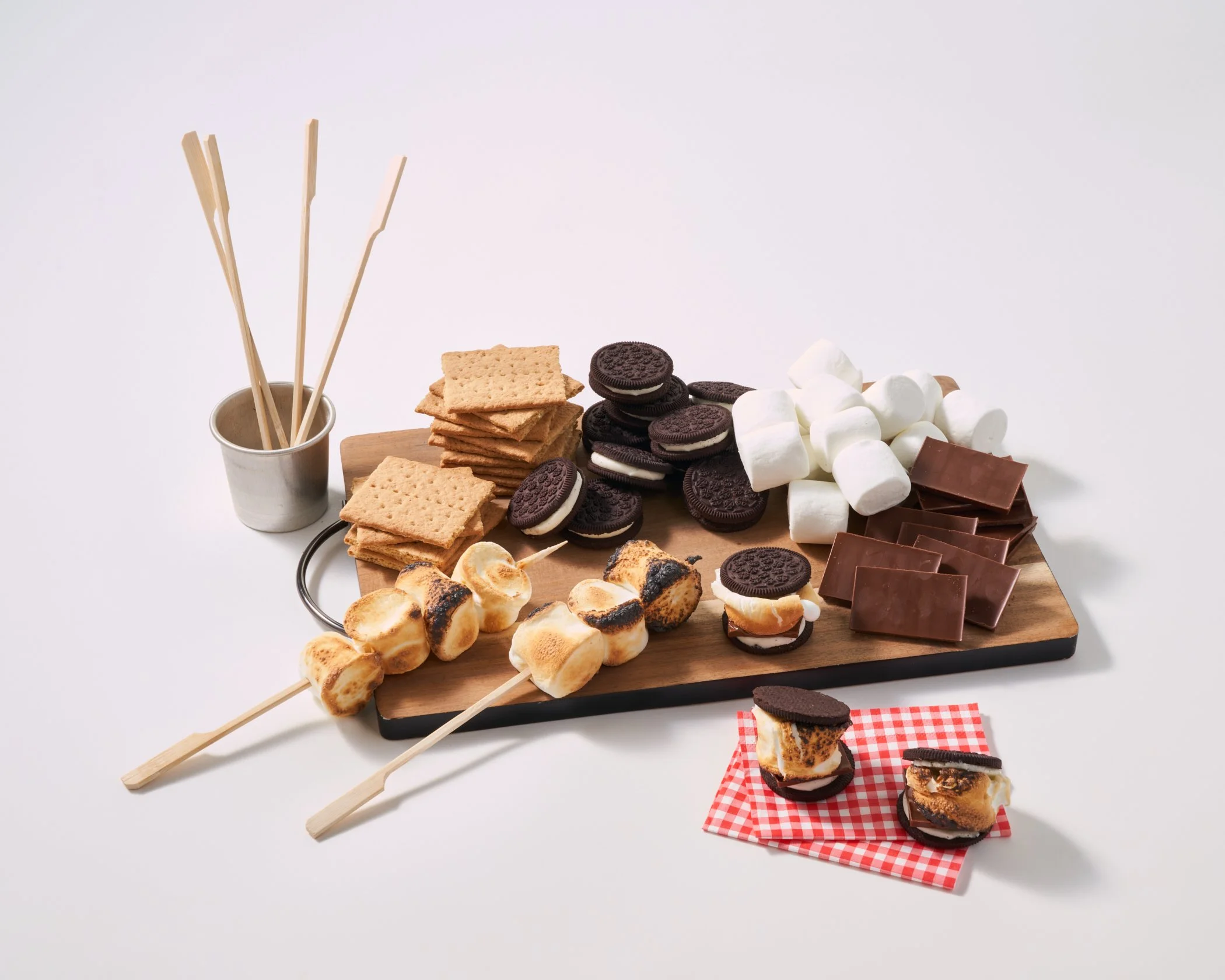 A wooden serving board with chocolate, vanilla, and s'mores desserts, marshmallows, cookies, graham crackers, and chocolate squares, with skewers and a small plate on a white background.