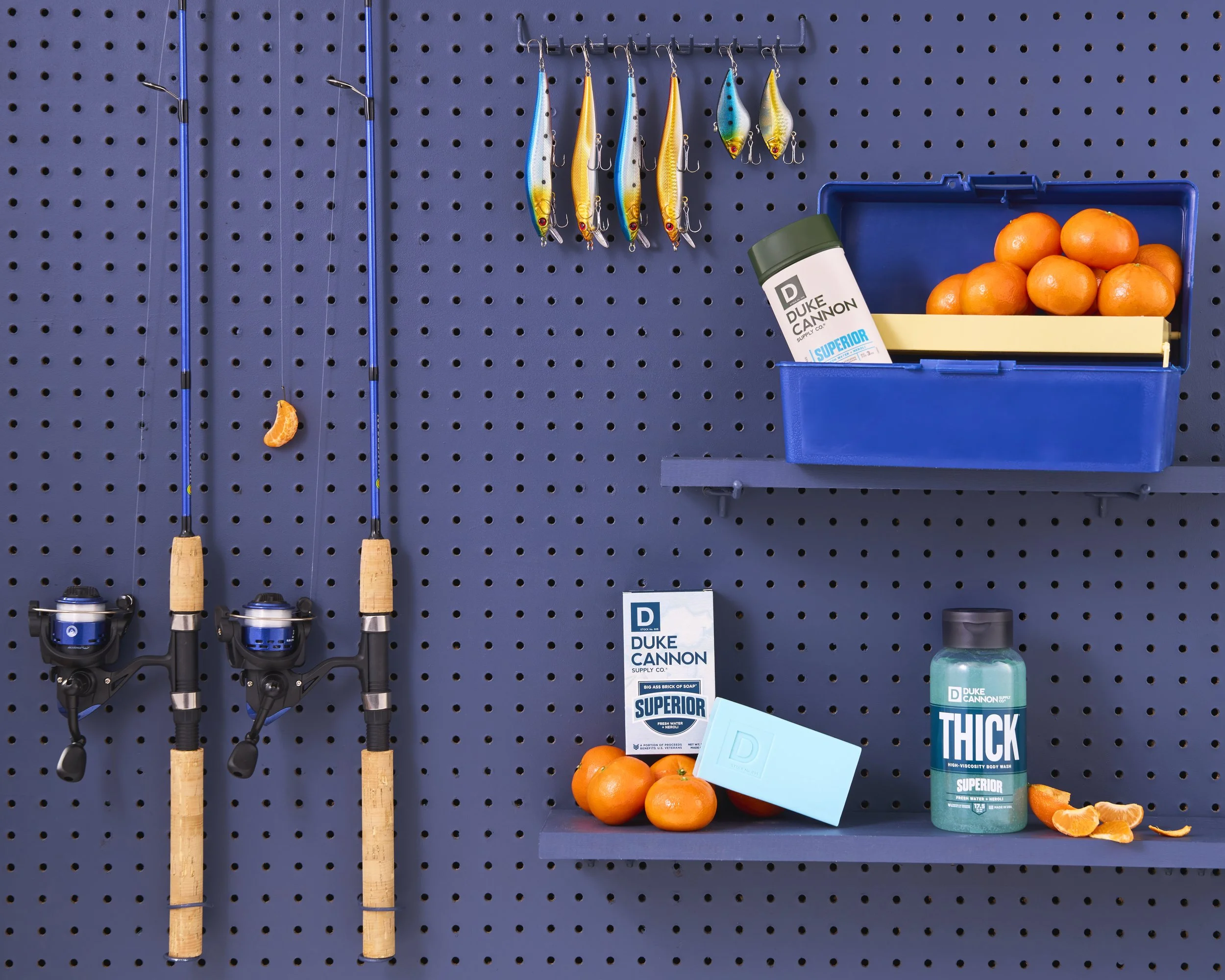 Fishing rods hanging on a blue pegboard, with a small orange peel, a blue plastic box containing oranges, a bottle of Duke Cannon soap, and a bar of Duke Cannon soap with oranges and orange peels nearby.