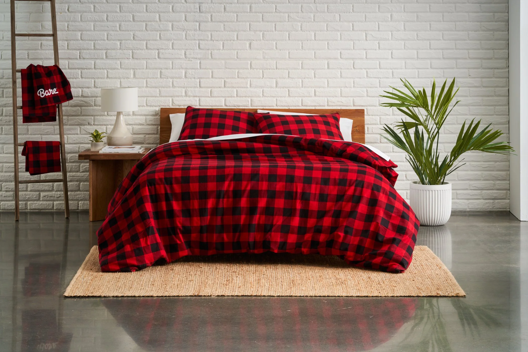 A bedroom with a white brick wall, wooden bed frame, and red and black plaid bedding. Next to the bed is a nightstand with books, a white lamp, and a small potted plant. On the left, a wooden ladder holds red and black plaid clothing, including a shi