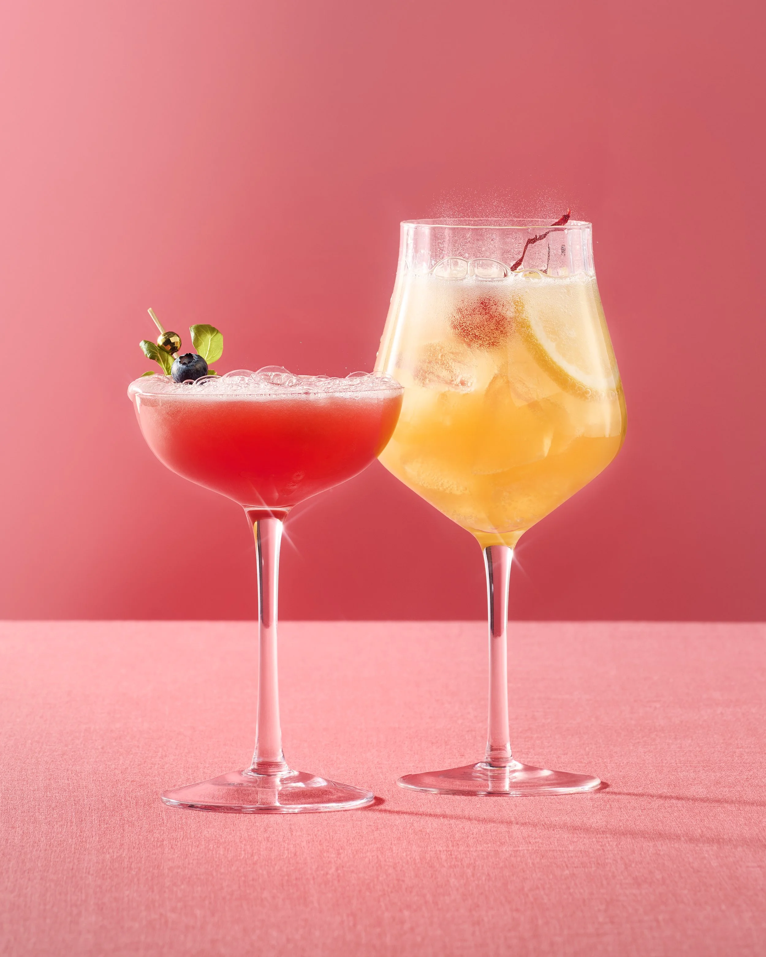 Two cocktail glasses, one with a pink drink garnished with berries, and one with a yellow drink with lemon slices, on a pink surface with a pink background.