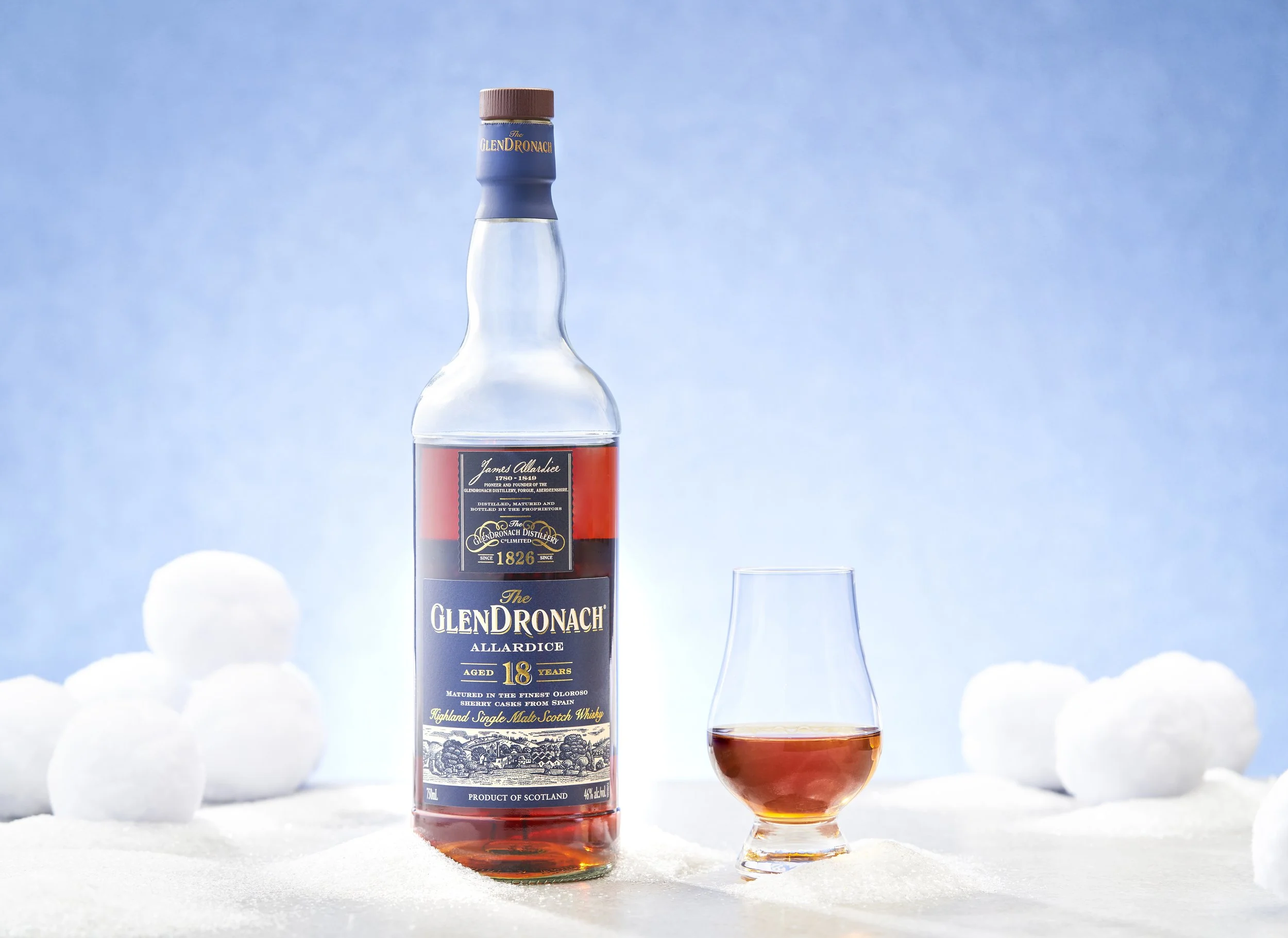 A bottle of GlenDronach 18-year-old single malt Scotch whisky with a glass of whisky beside it on a snowy surface, with snowballs in the background and a light blue sky.