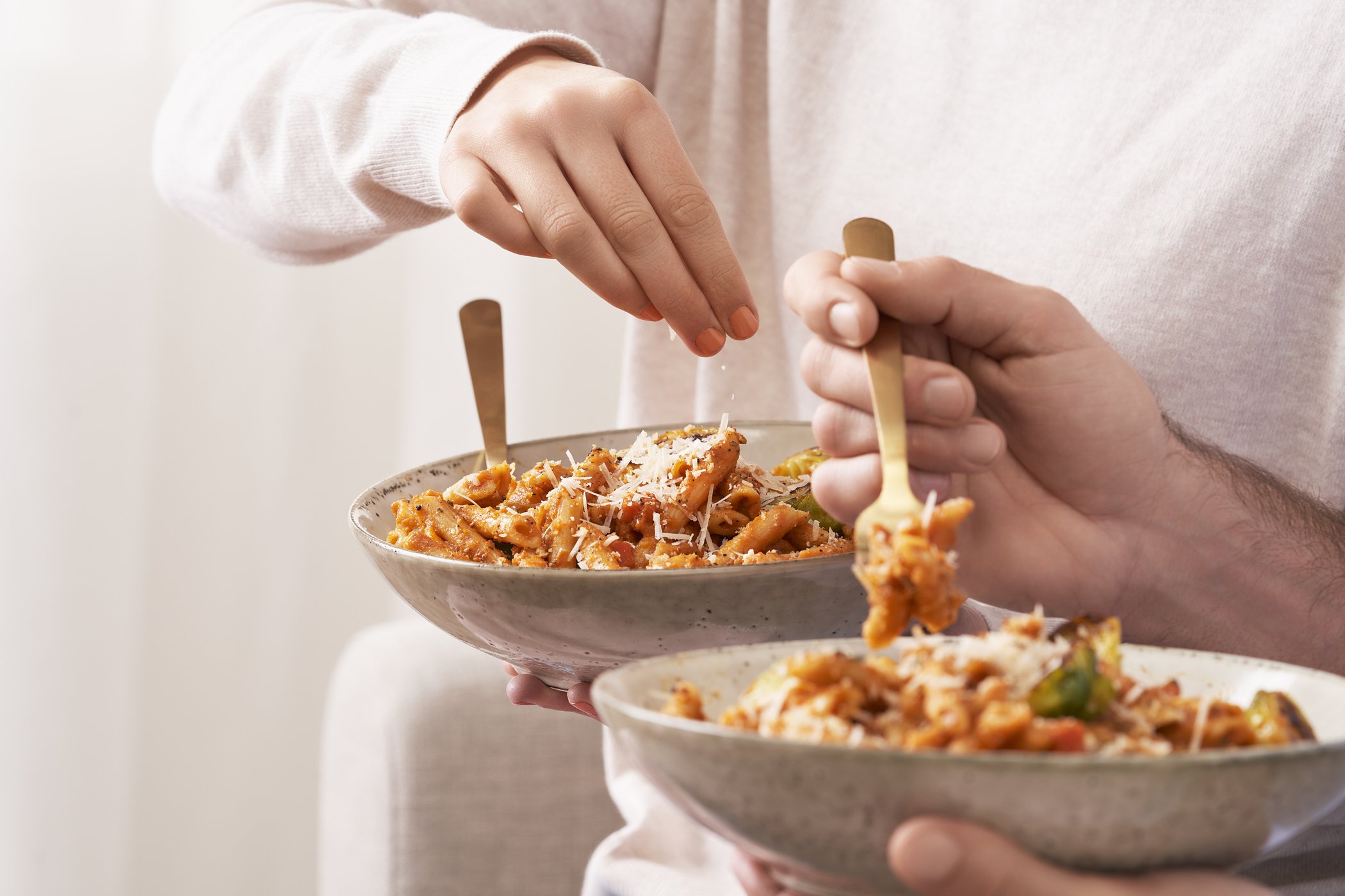 Two people sharing bowls of pasta with shredded cheese, with one person sprinkling cheese over their bowl.