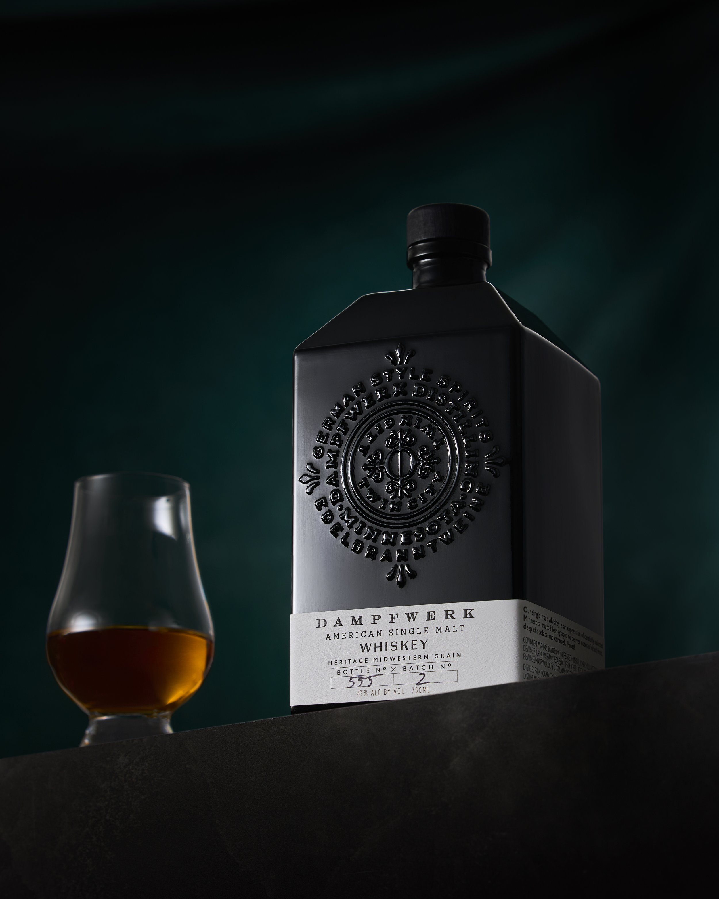 A black bottle of Dampfwirk American single malt whiskey with a white label, next to a glass of amber whiskey on a dark surface against a dark background.