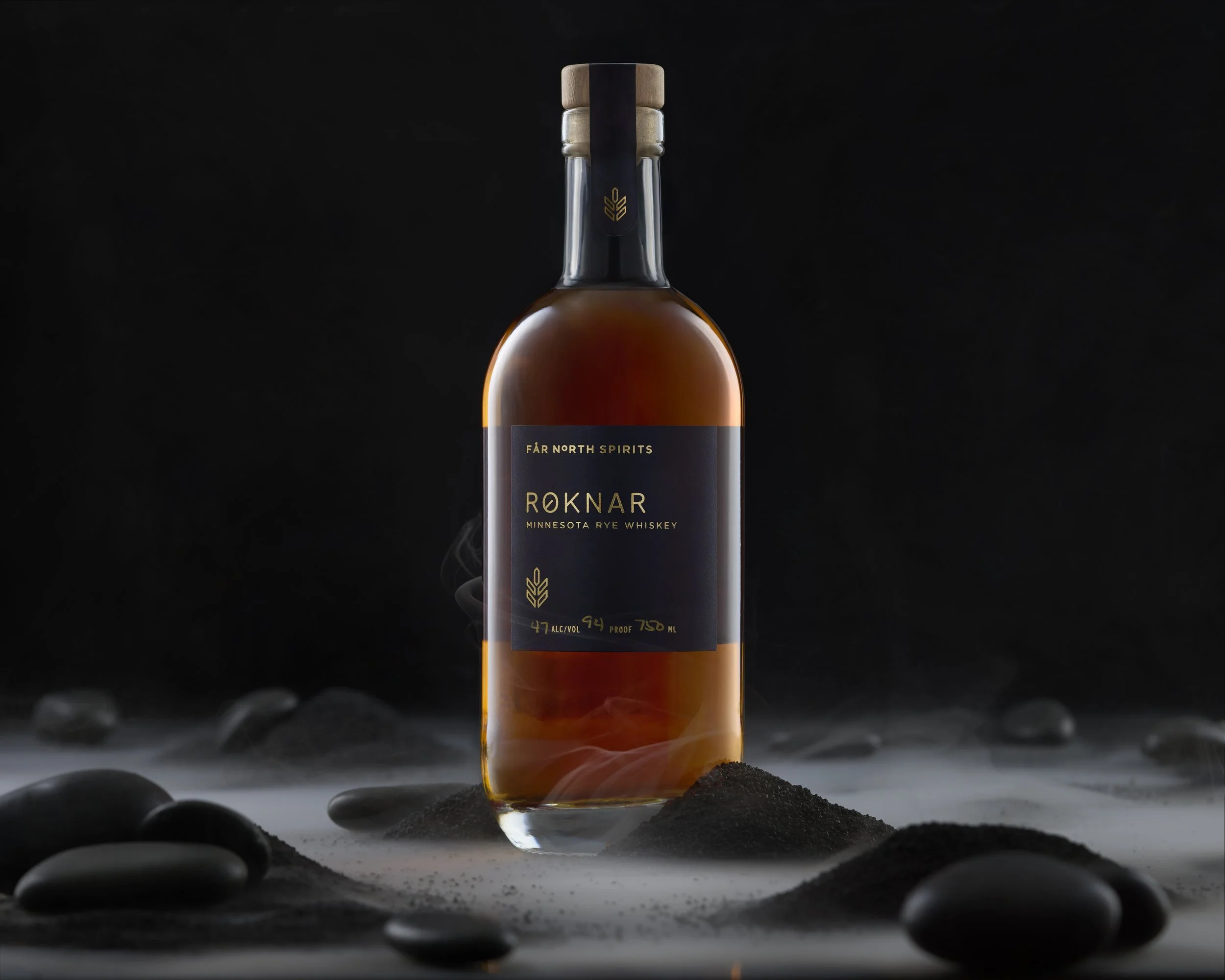 A bottle of Røknar Minnesota rye whiskey standing upright on a surface scattered with black stones and sand, with a dark background and subtle steam rising around it.