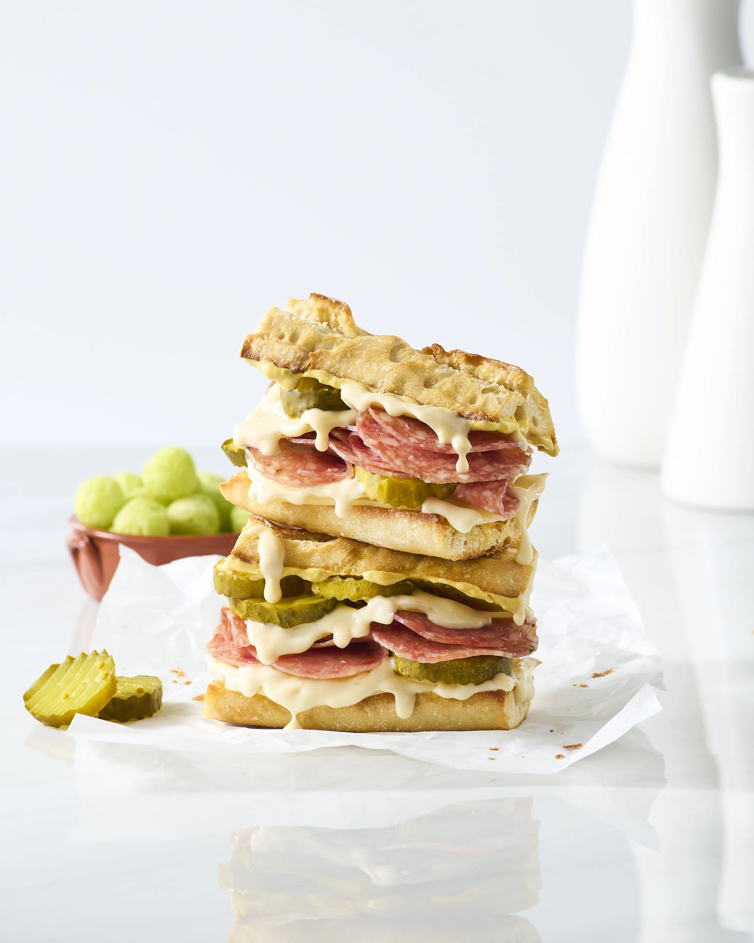 Stacked sandwiches with pickles, melted cheese, and deli meats on white paper, with a bowl of green balls in the background.