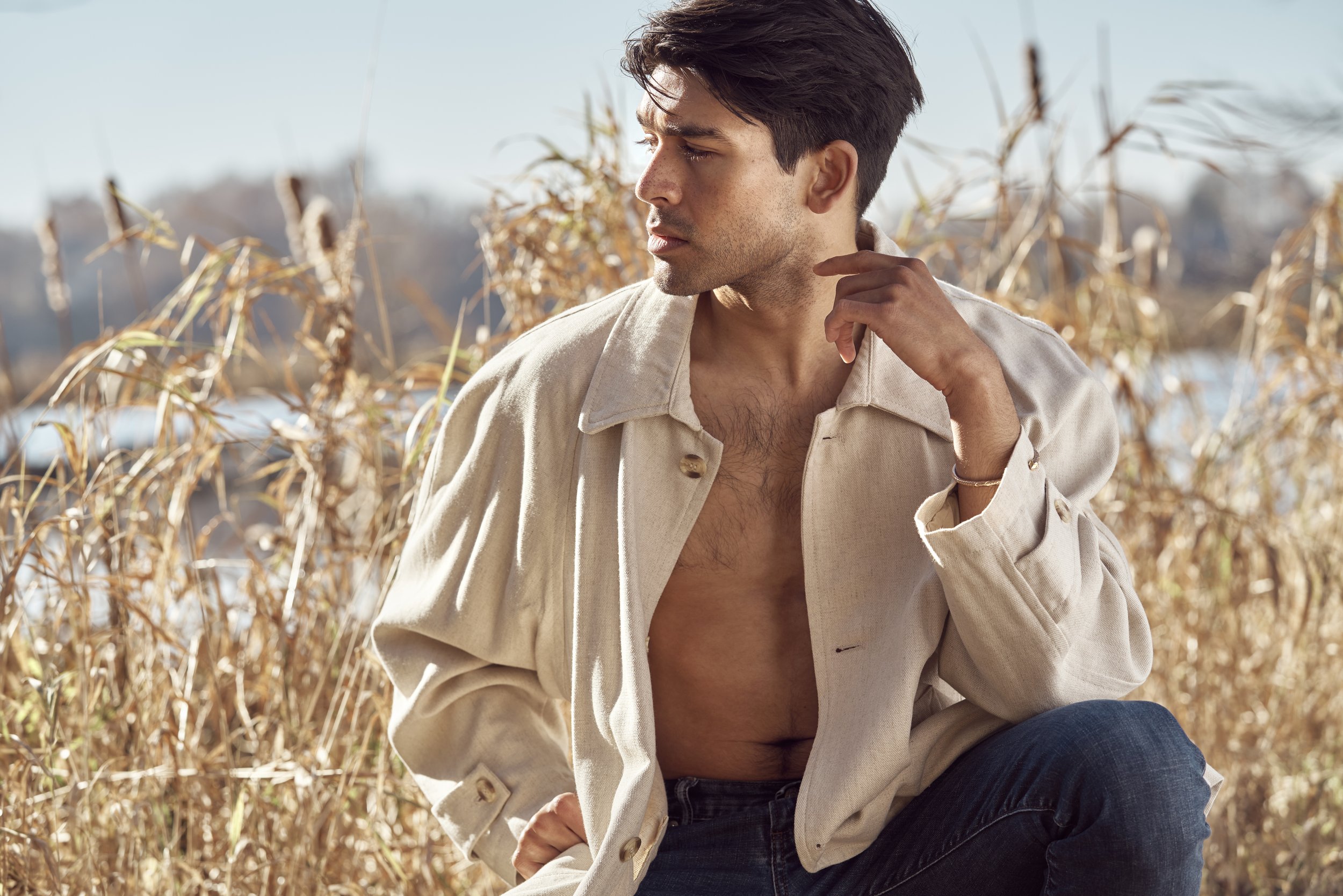 A young man in a beige jacket, with the jacket partially open to reveal his bare chest, sitting outdoors in a field of dry grass with water in the background, looking to the side.