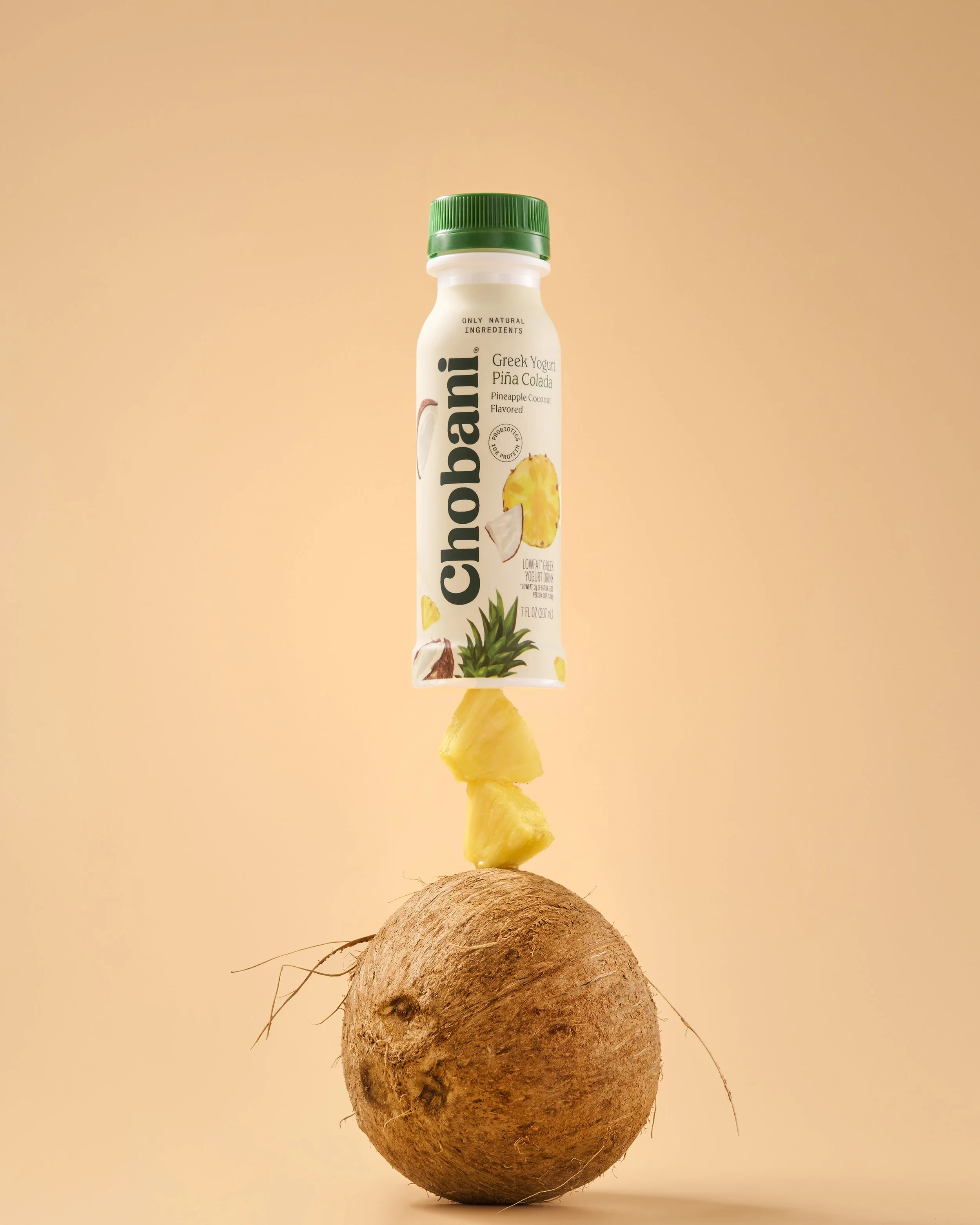 A chocolate Chobani yogurt bottle suspended above a whole yam with pineapple pieces between them, against a peach-colored background.