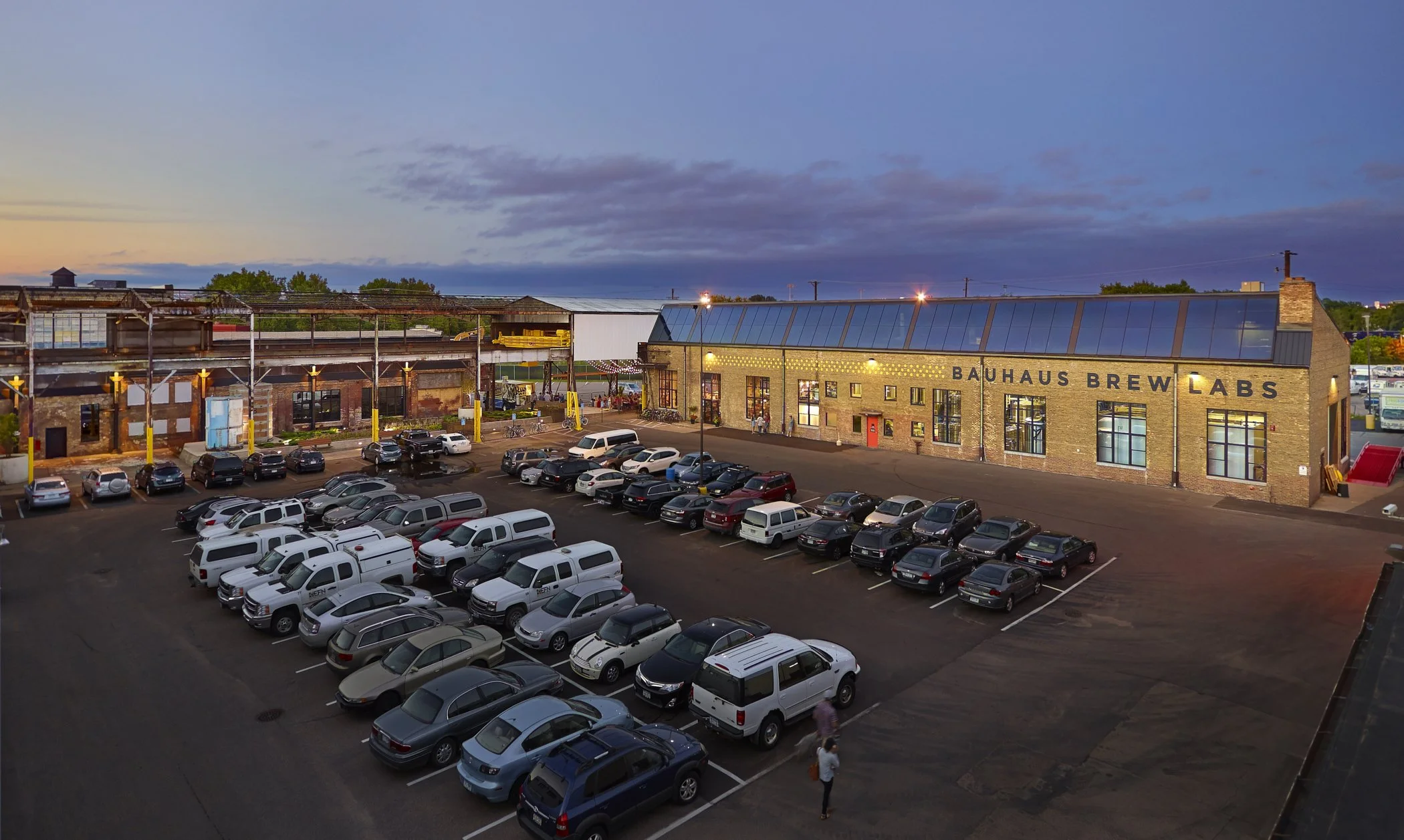 Parking lot with various cars in front of Bauhaus Brew Labs building, urban area during evening with sunset sky, some people walking, industrial style building with large windows and signage, distant trees and sky