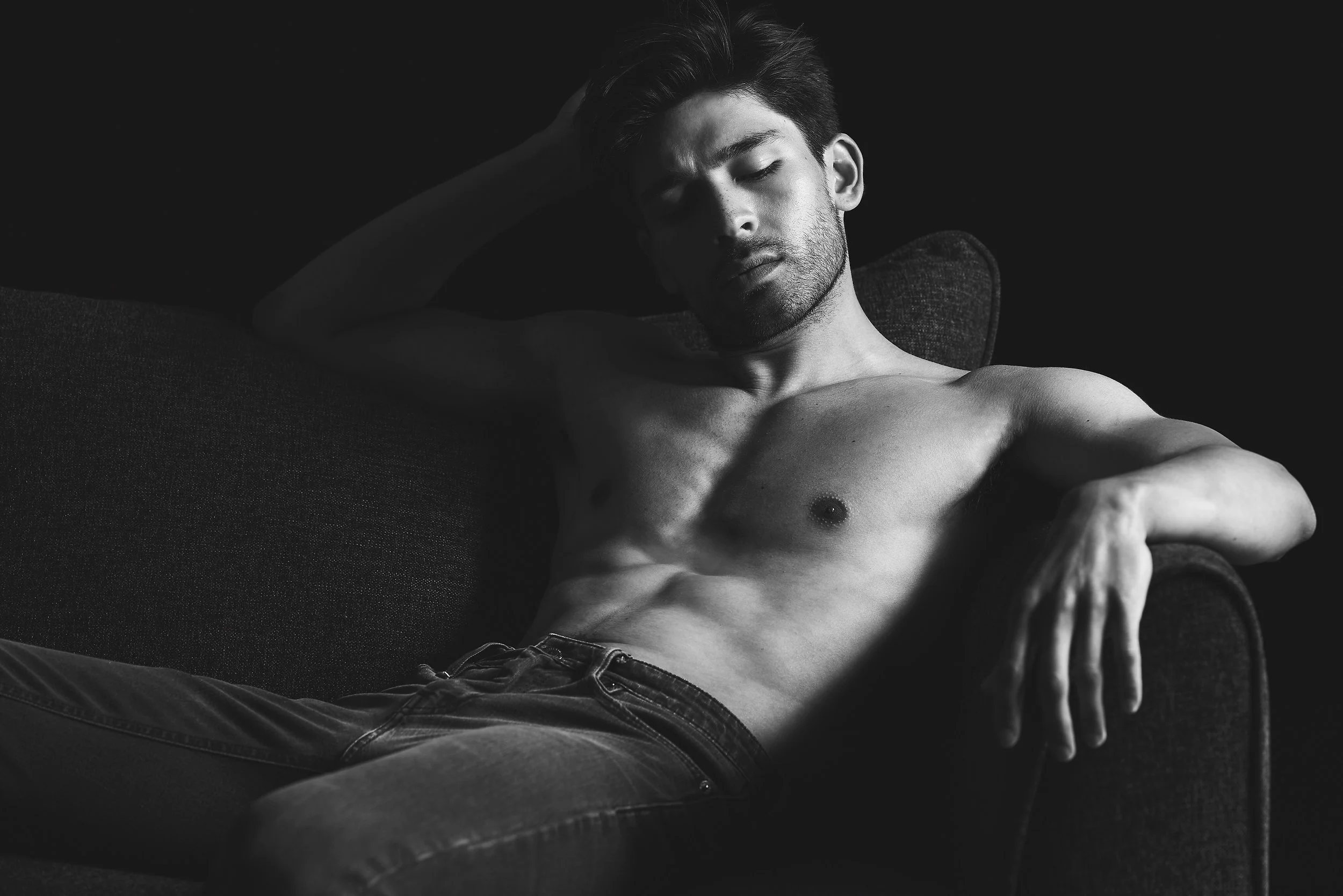 A shirtless young man with dark hair and light skin reclining on a dark sofa against a dark background, with his eyes closed.