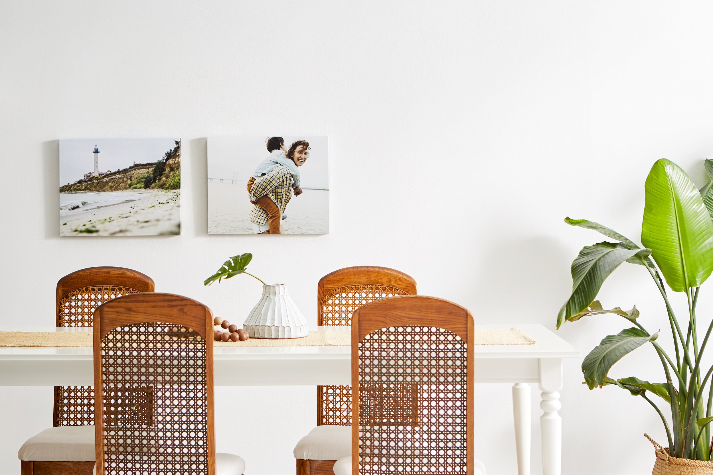 A bright dining room with a white table, four wooden chairs with cane backs, a white vase with a green leaf, and a large green plant. Two framed photos hang on a white wall behind the table.