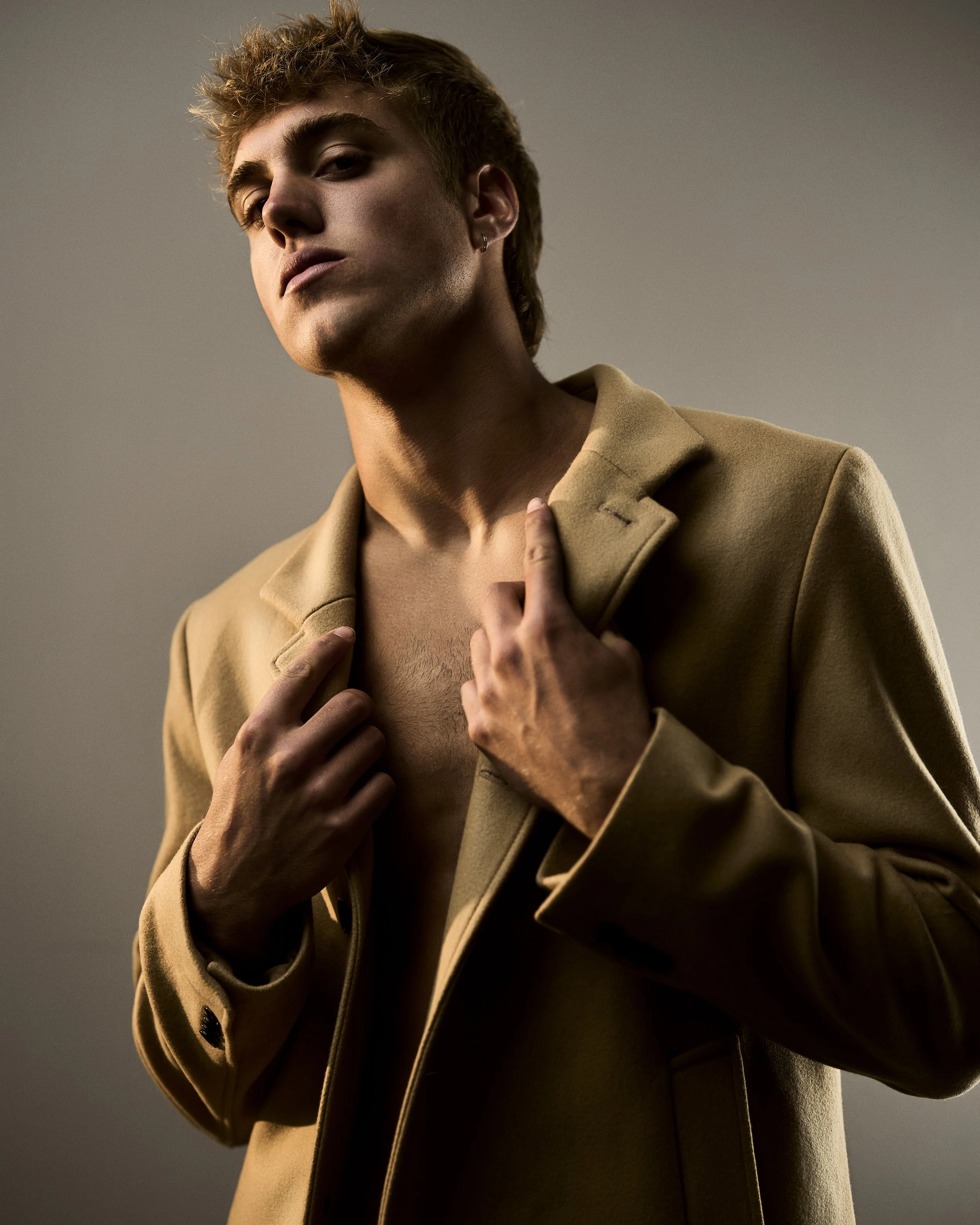 A young man with curly hair and earrings wearing a beige coat, posing with his hand touching his chest against a plain background.