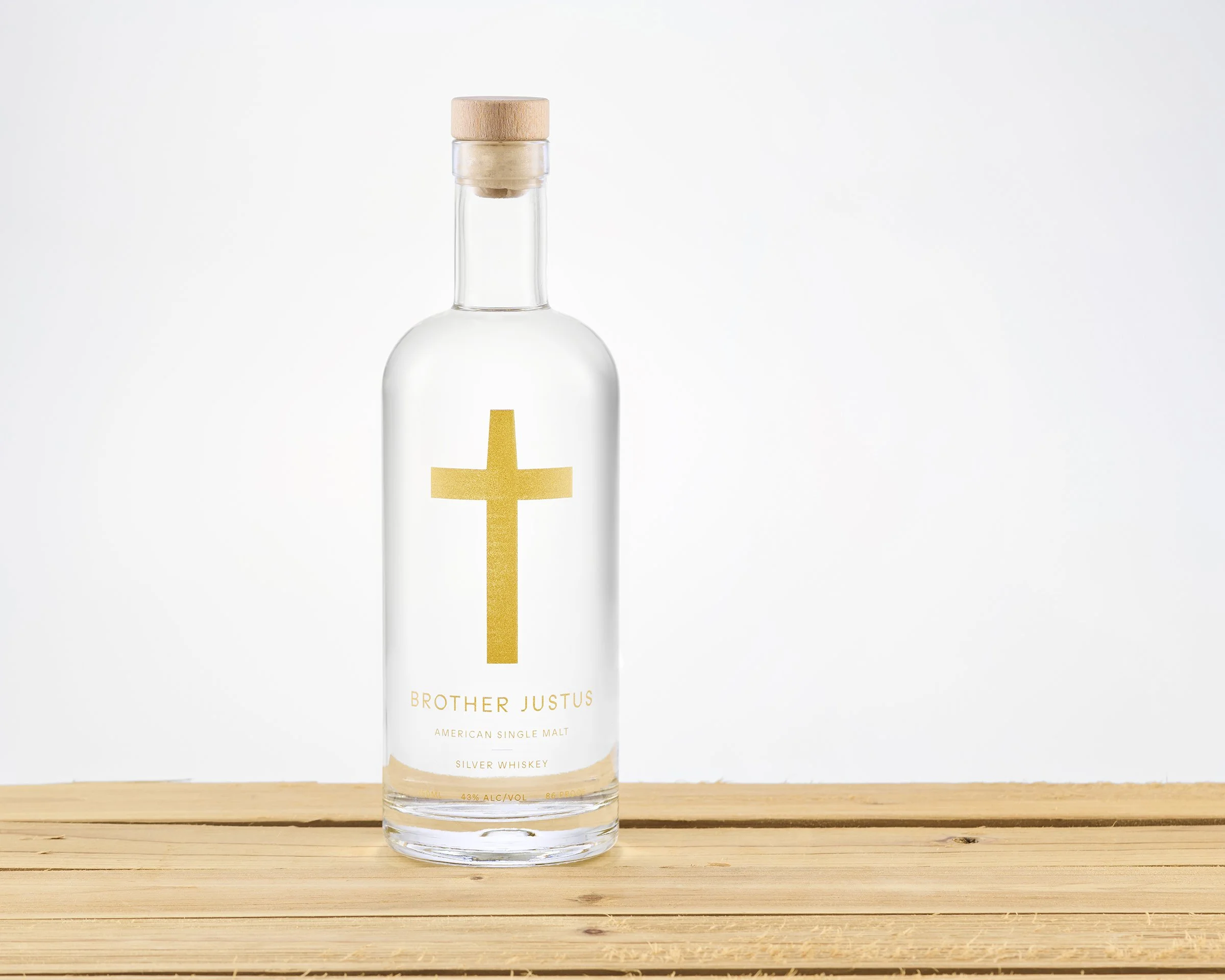 A clear glass bottle of Brother Justus American single malt silver whiskey with a gold cross label, standing on a wooden surface, against a plain light background.