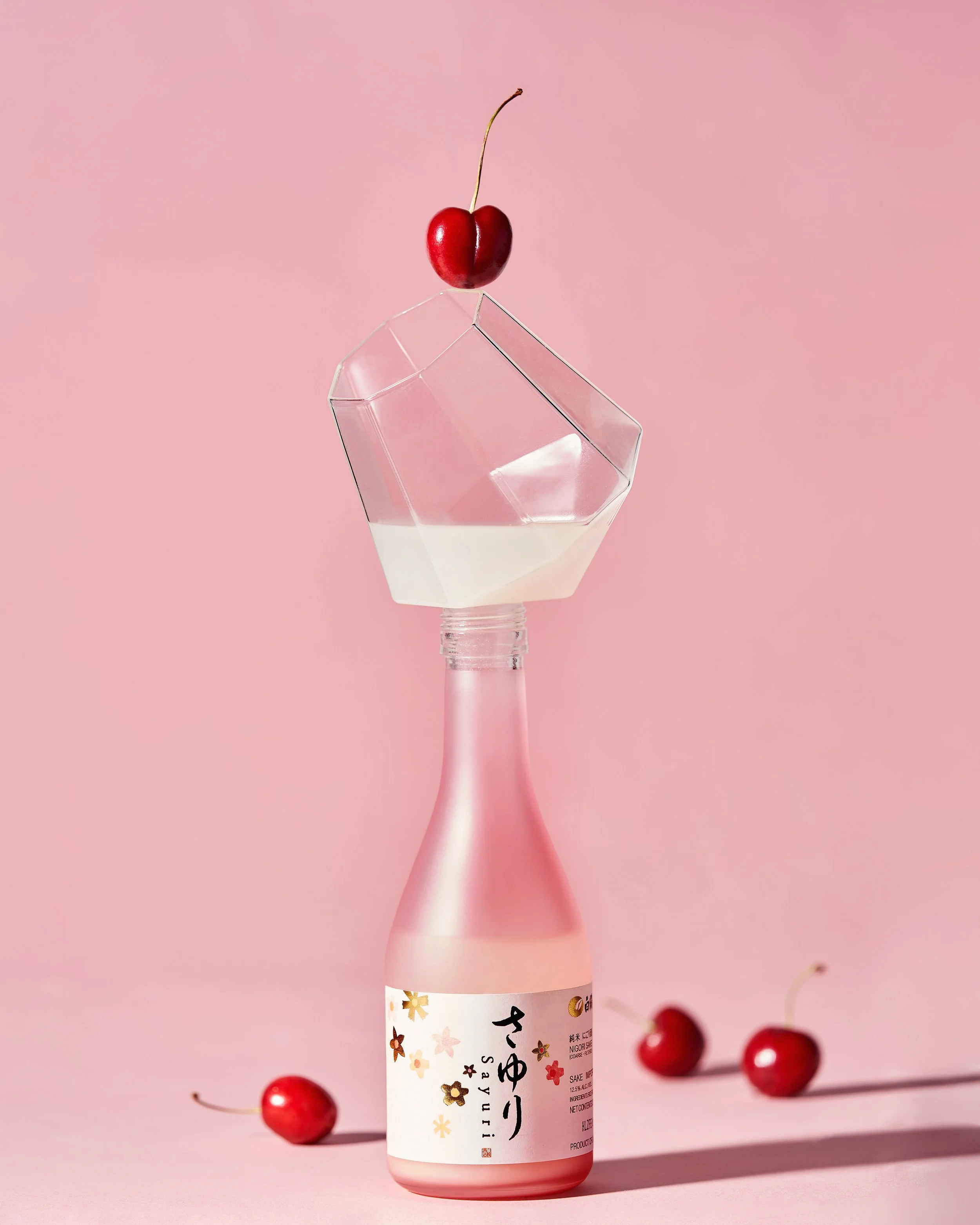 A pink bottle with a Japanese label, a clear glass container with some liquid, a cherry on top of the container, and three cherries around the bottle on a pink background.