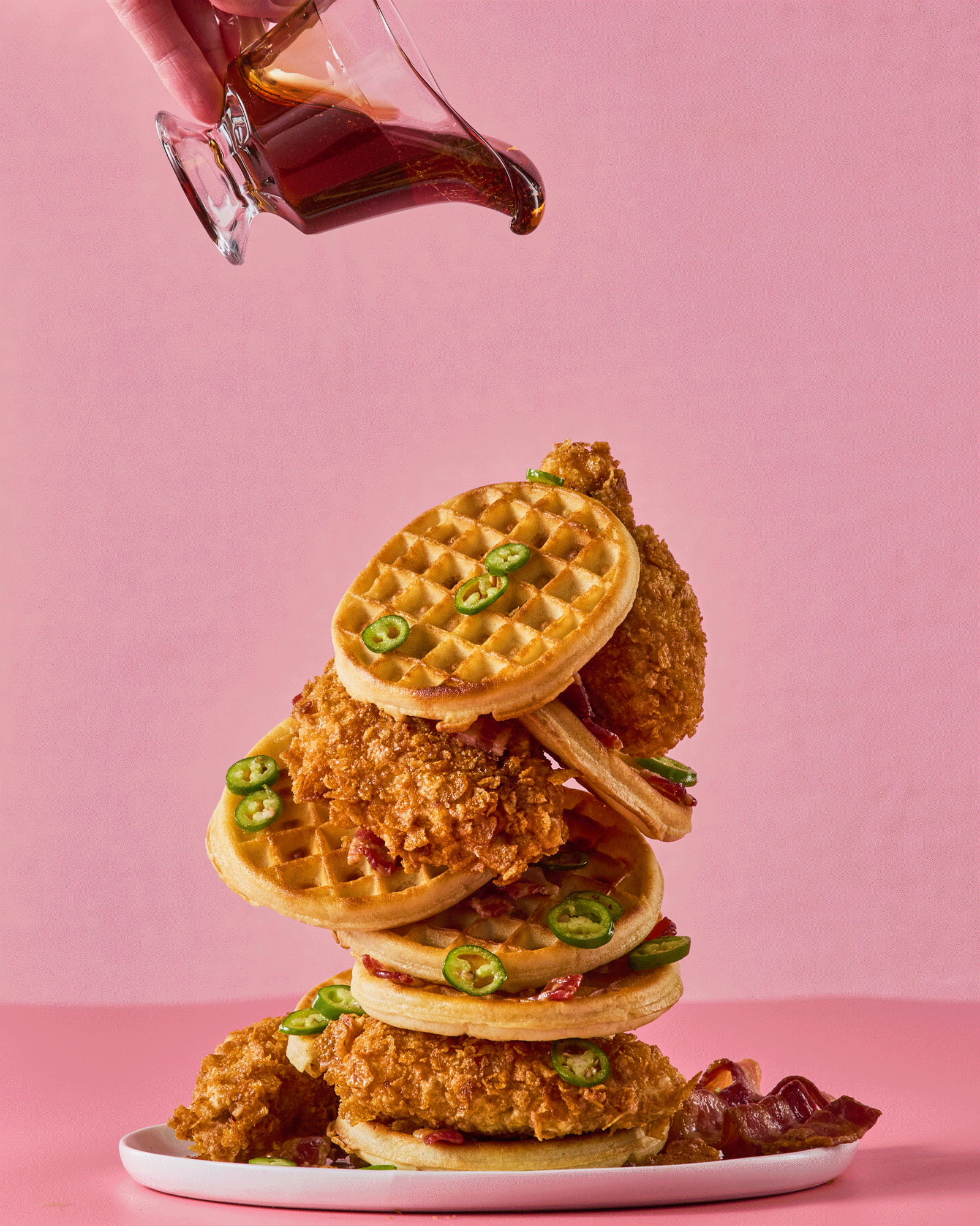 Syrup pouring over stack of chicken and waffles on a pink surface and background.