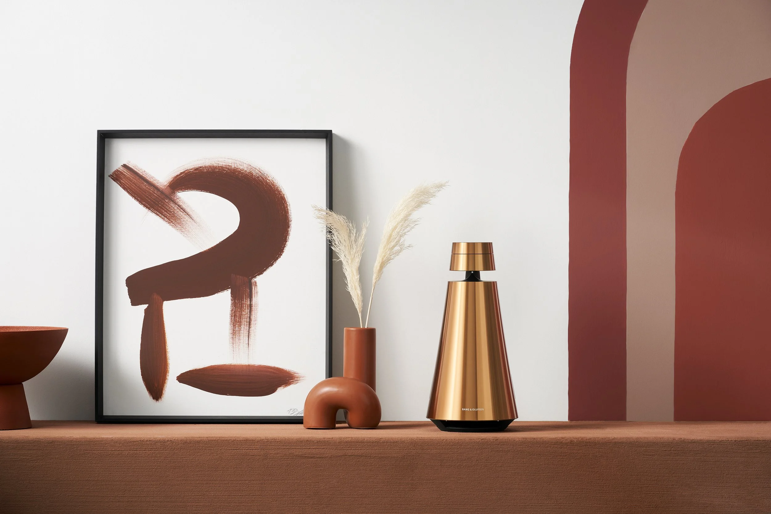 Decorative interior scene with a framed abstract art print, a vase with dried pampas grass, a metallic lamp, and a terracotta-colored vase on a wooden surface against a white wall with red painted accents.