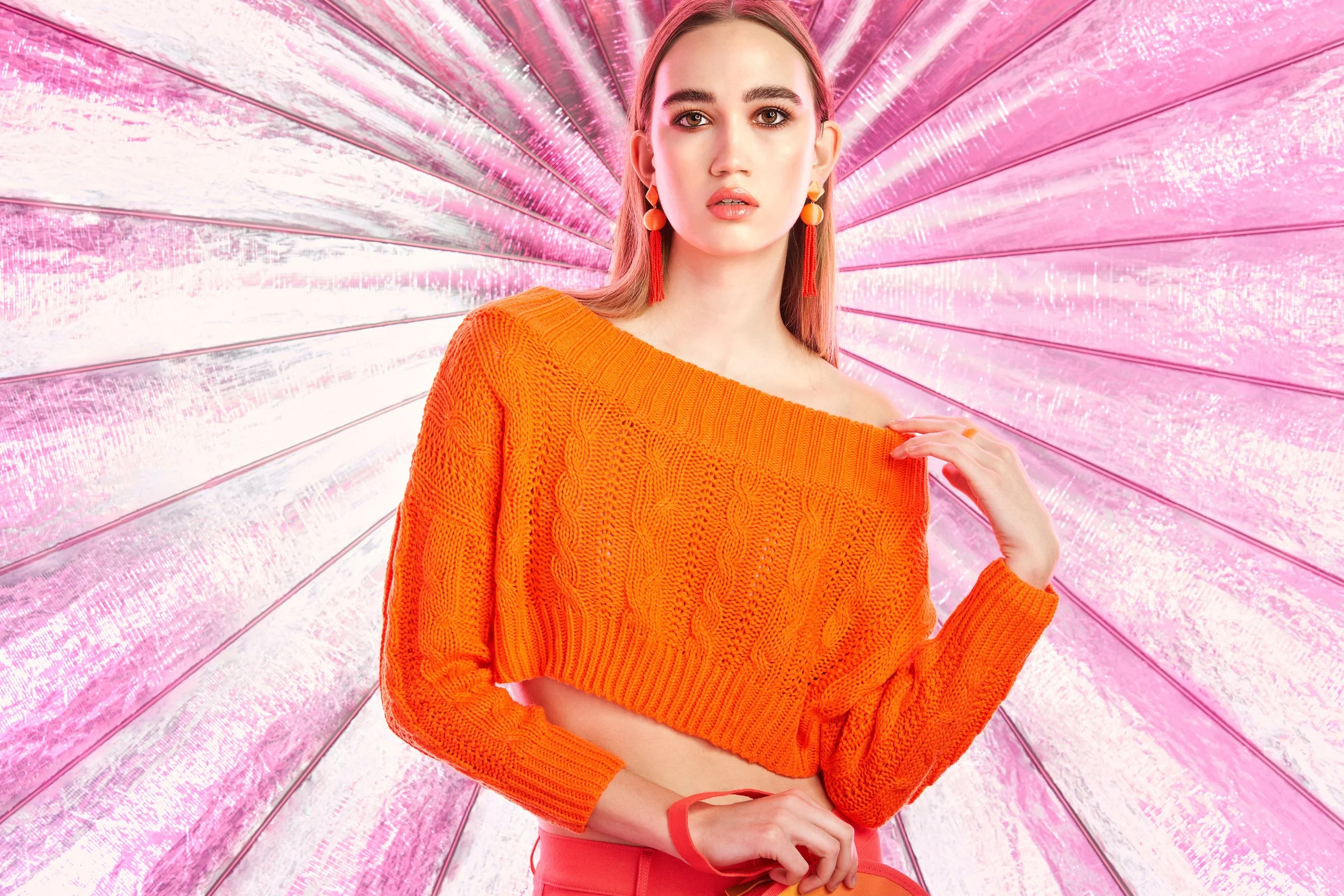 A young woman with light brown hair and wearing orange earrings and an orange sweater, standing in front of a pink reflective background with a radiant pattern.