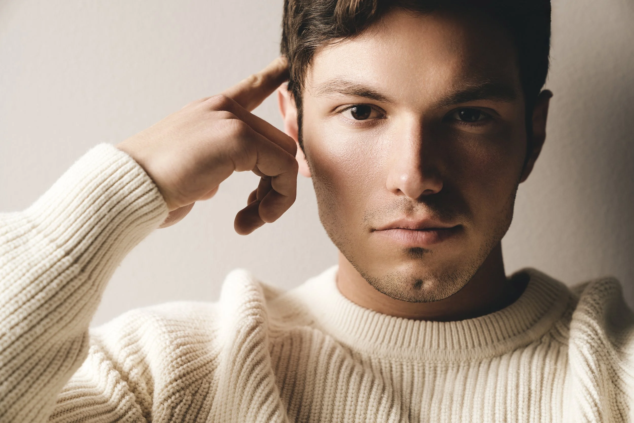 Close-up of a young man with light brown skin, short dark hair, wearing a white knit sweater, touching his temple with his right hand, and looking directly into the camera against a plain, light background.