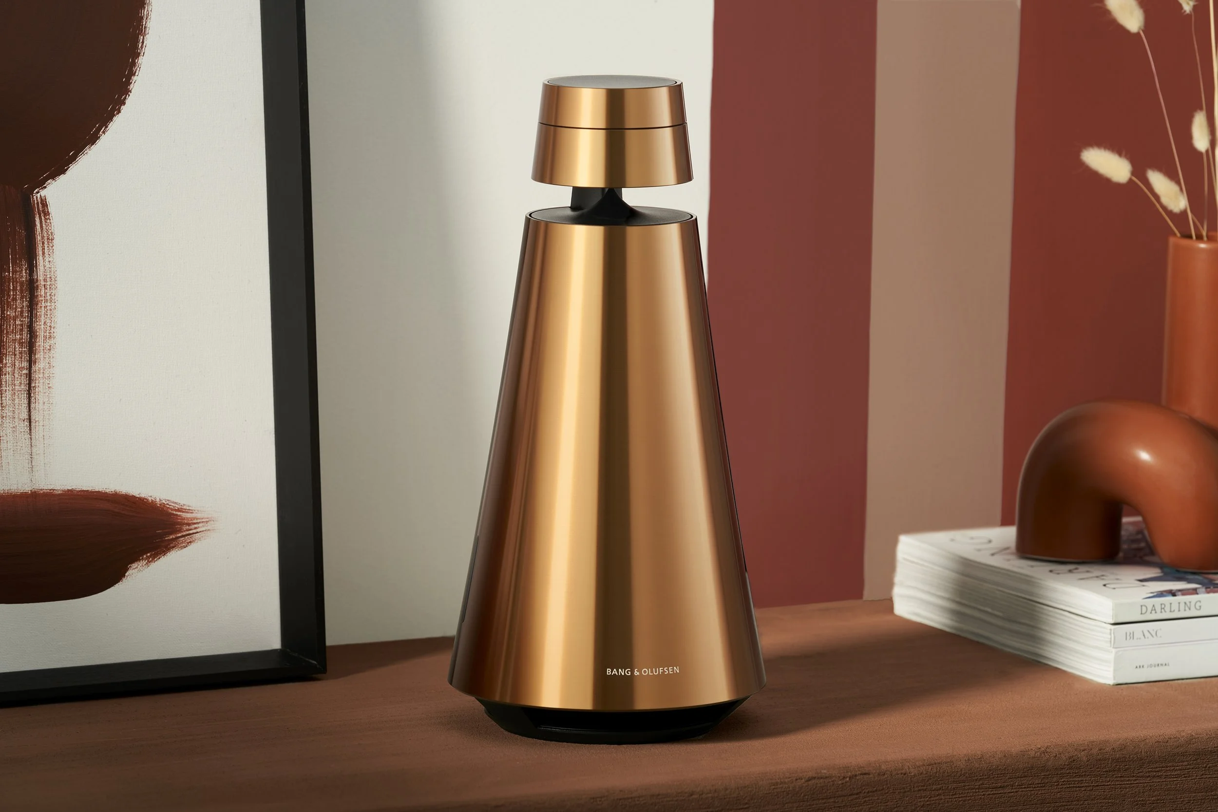 A copper-colored Bang & Olufsen speaker on a wooden surface, with a stack of white books labeled 'DARLING' and 'BLANC' to its right, and an orange vase with white dried flowers in the background.
