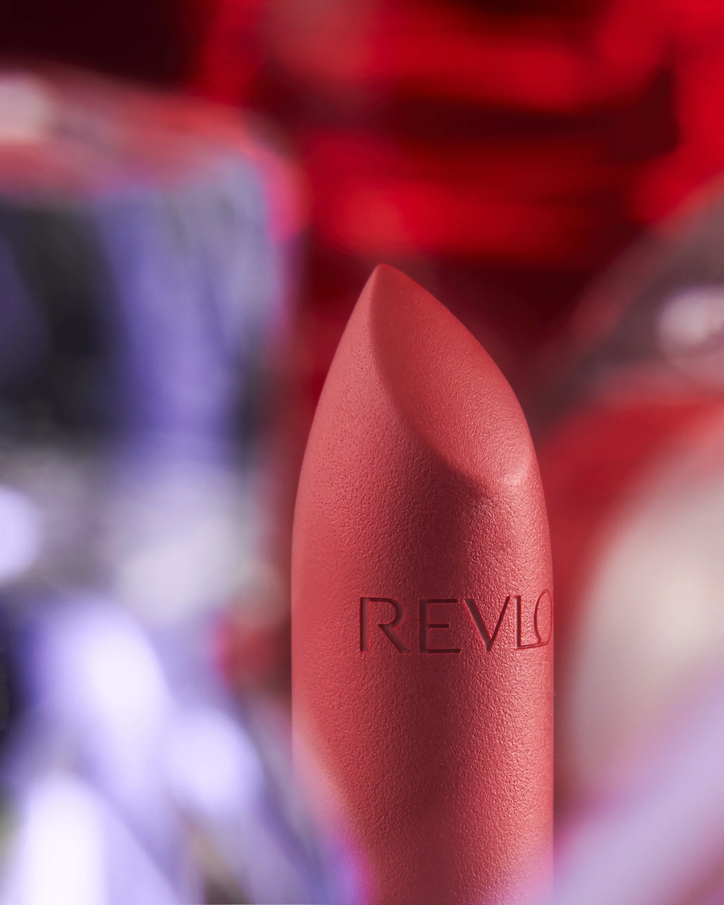 Close-up image of a coral-colored lipstick with the brand name 'Revlon' embossed on it. The background is blurry with hints of red, purple, and white colors.