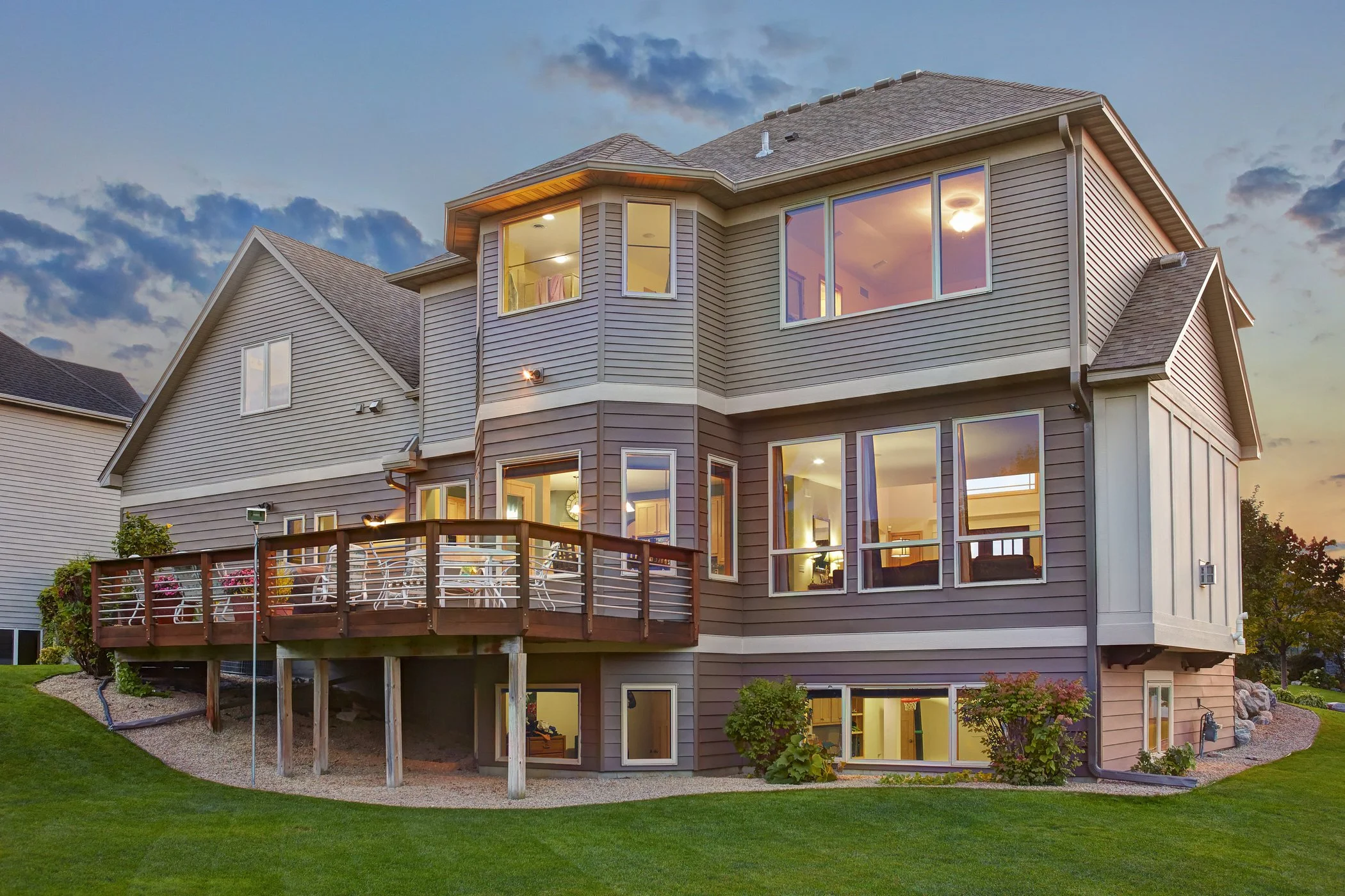 A two-story house with gray siding, multiple large windows, a wooden deck with chairs and table, and a green lawn at sunset.