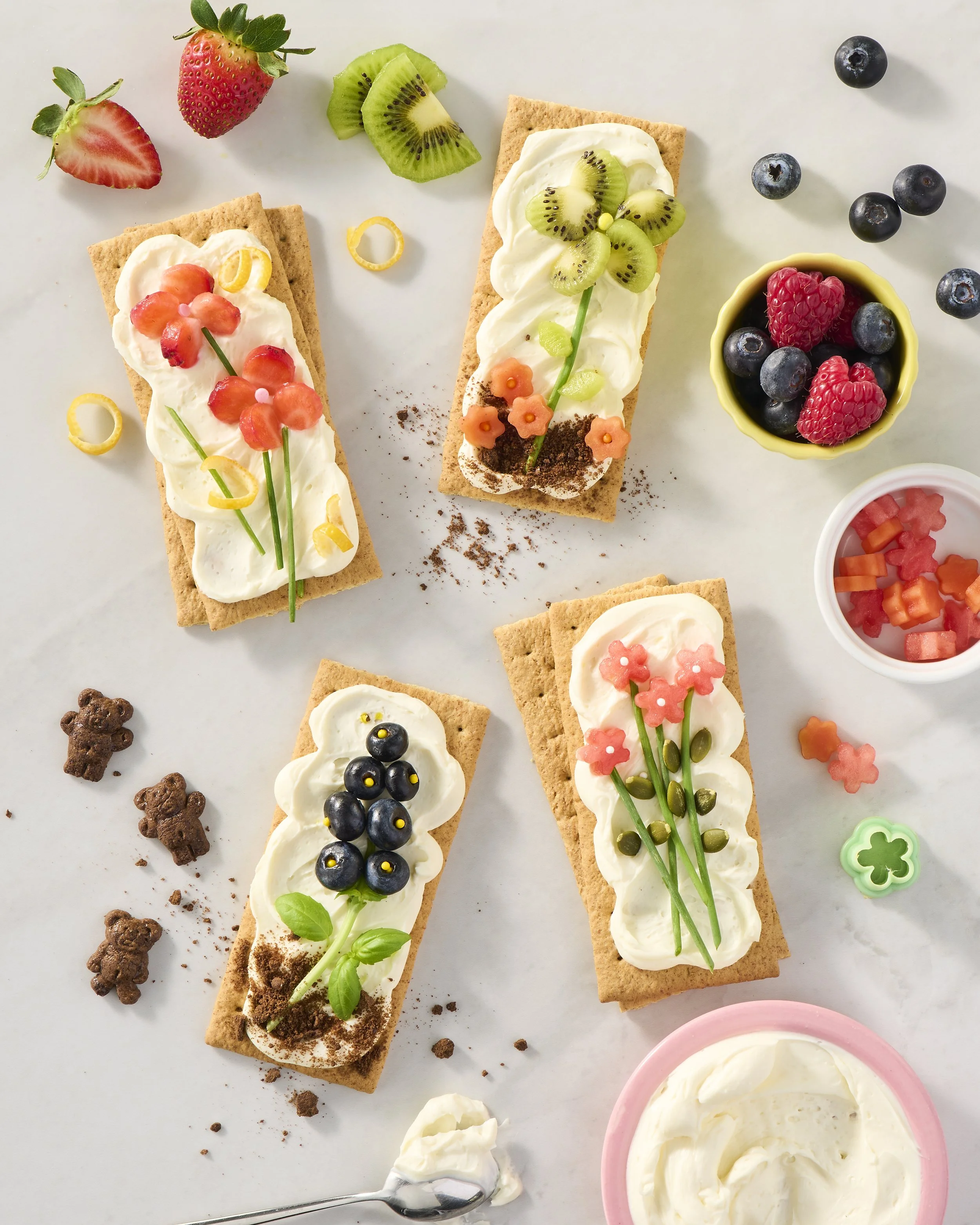 Sweet crackers topped with whipped cream, berries, and decorative toppings arranged on a white surface with scattered berries and small bear-shaped cookies.