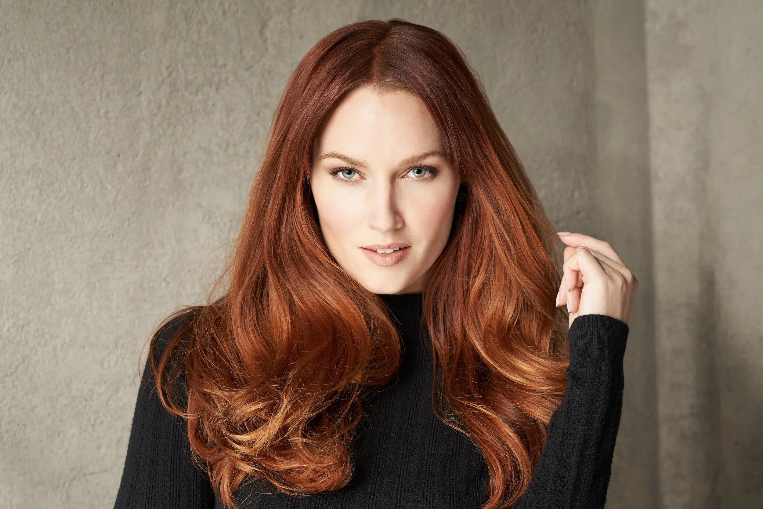 Woman with long, wavy red hair wearing a black top against a neutral background.