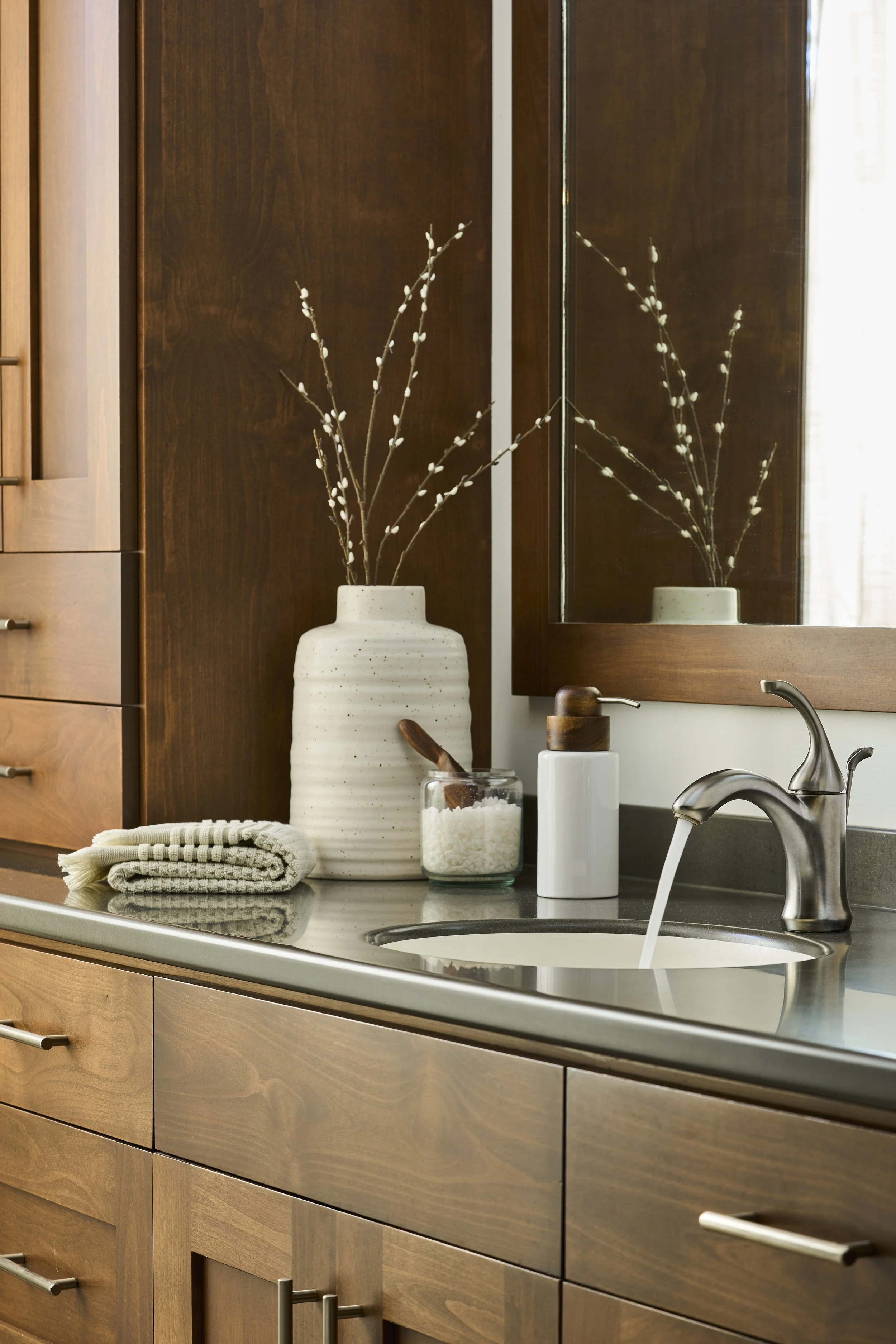 Bathroom vanity with a large beige vase holding dried branches, a small rolled towel, soap dispenser, container of bath salts, and a mirror reflecting the vase.