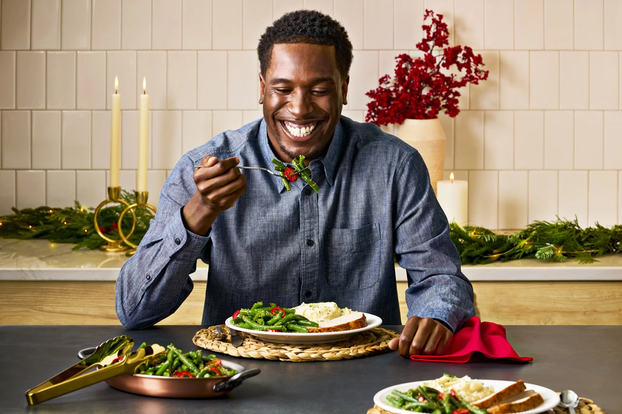 A man happily eating a holiday meal with green beans, mashed potatoes, and bread in a decorated kitchen with candles and red flowers.