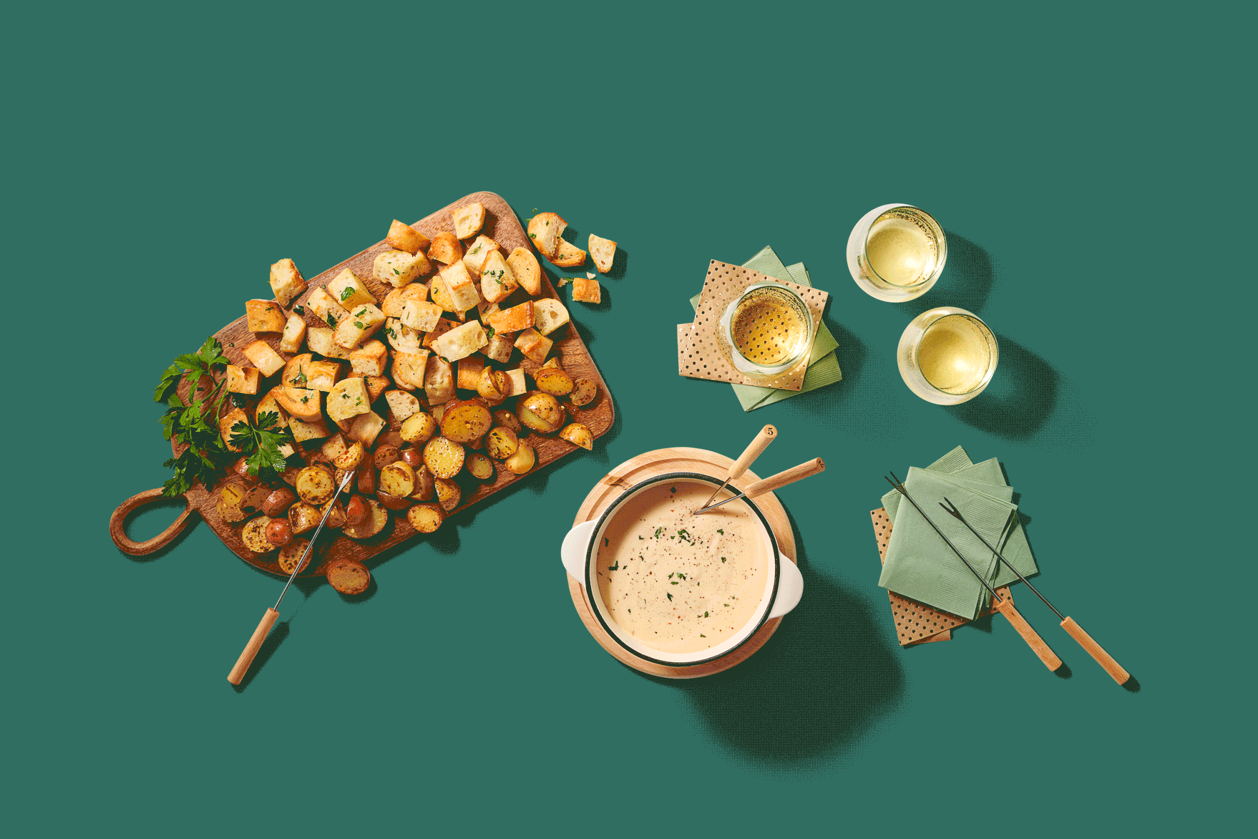 Overhead view of a table with toasted bread and roasted potatoes on a wooden serving board, two glasses of white wine, a bowl of creamy soup with chopsticks, and green napkins.