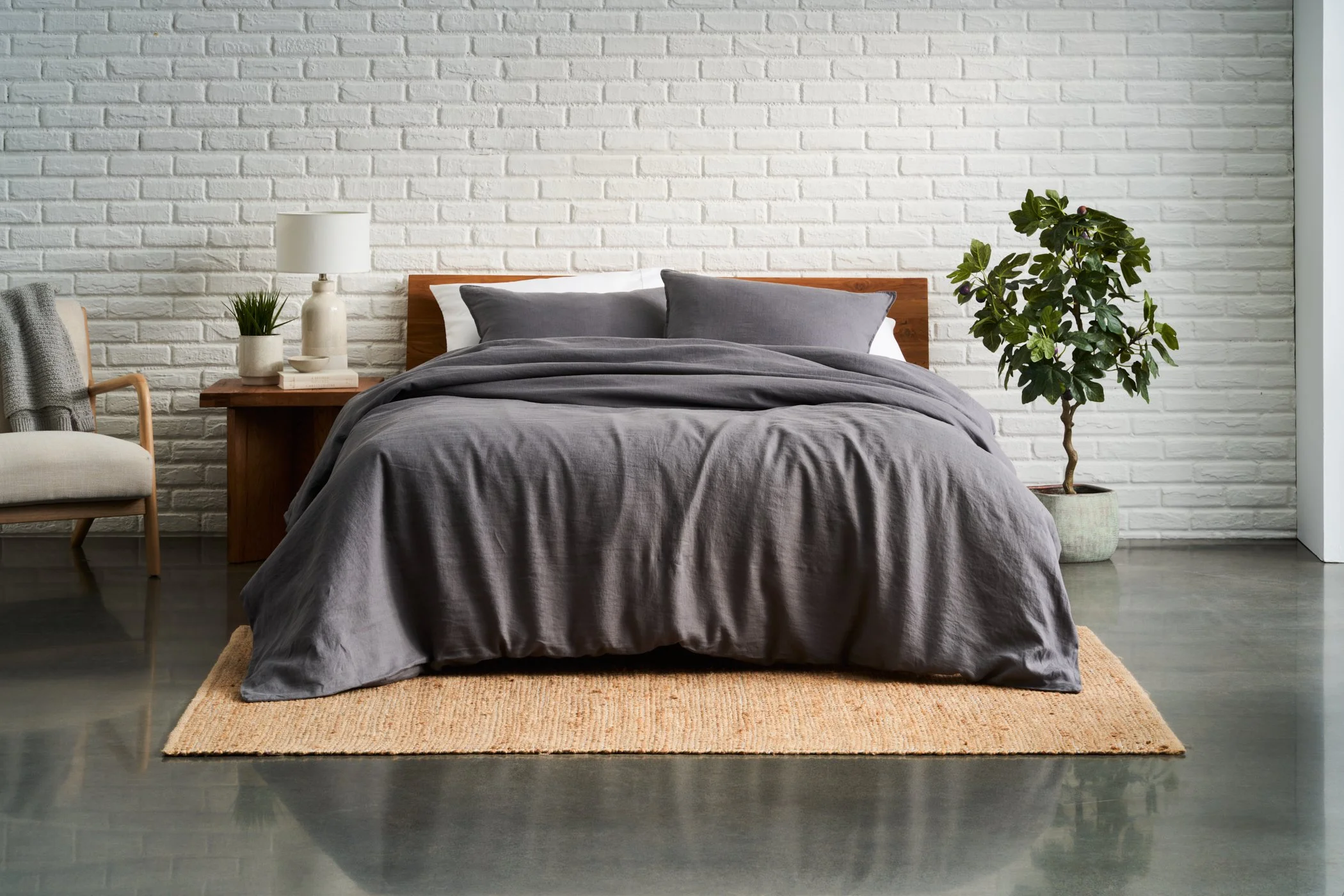 A bedroom with a bed covered in gray bedding, two gray pillows, a nightstand with a lamp, some books, and a plant on top. There is a chair draped with a gray blanket and a large green plant in a pot next to the bed. The background features a white br