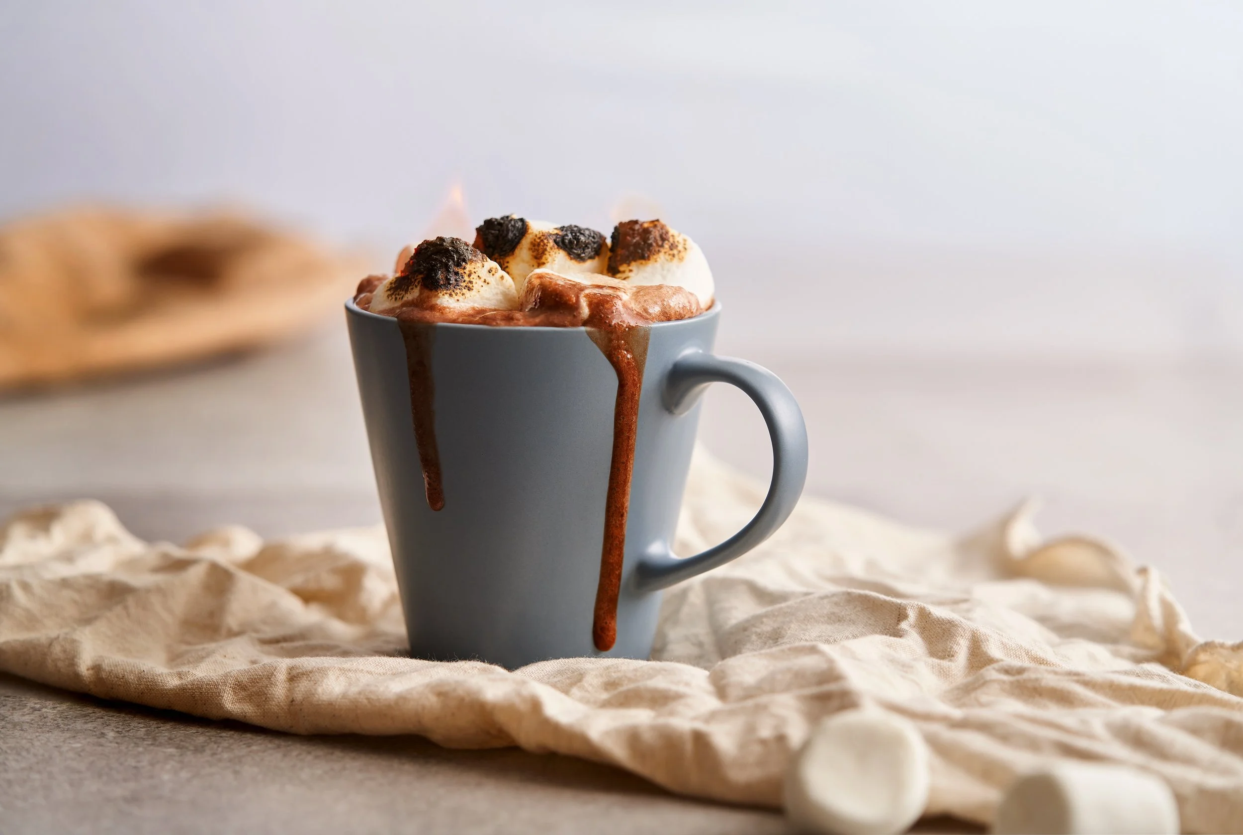 Gray ceramic mug filled with hot chocolate topped with marshmallows and chocolate crumbles, with chocolate syrup dripping down the side, on a beige cloth with a plate of cookies in the background.