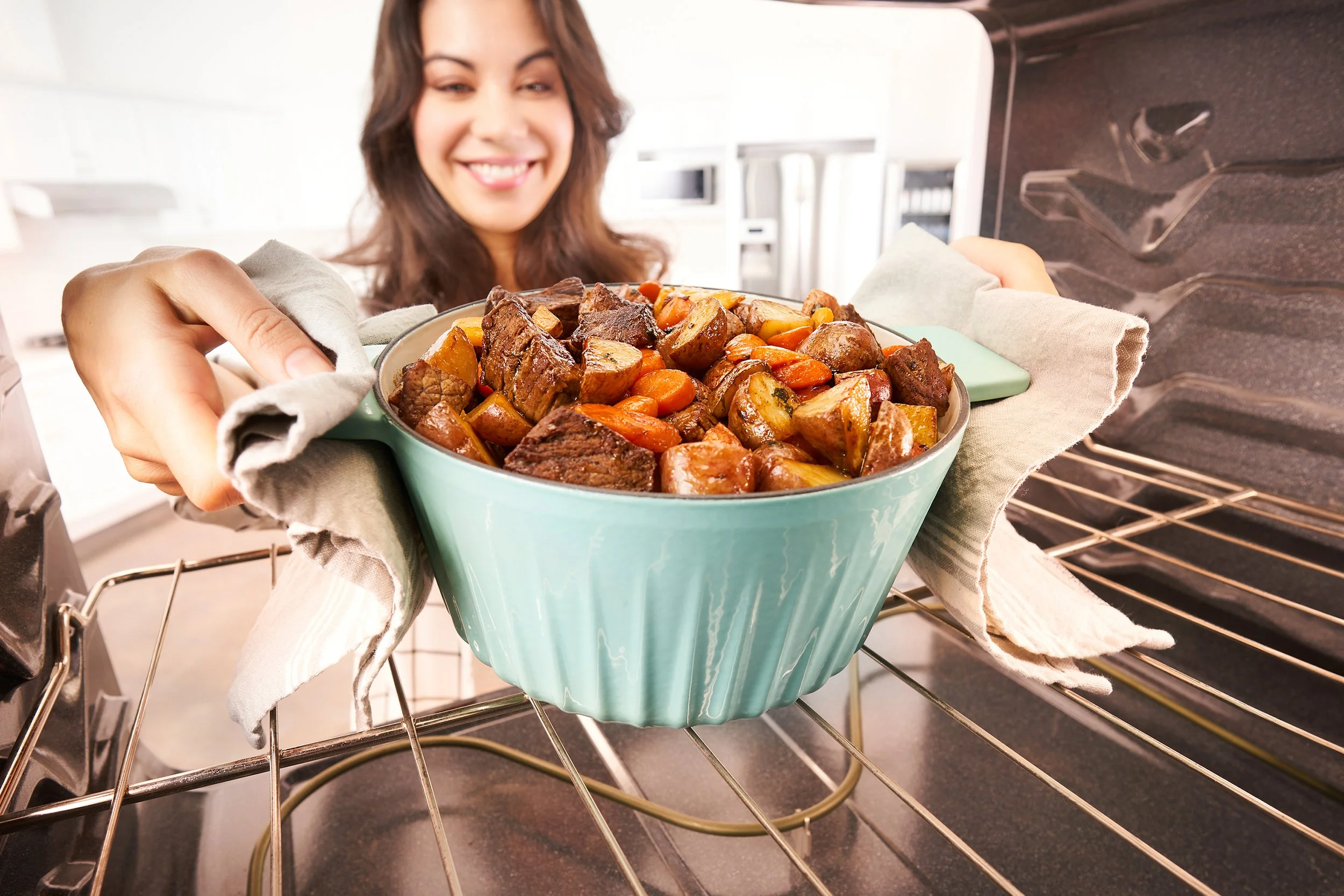 A woman putting a dish of cooked beef stew and vegetables into an oven, smiling at the camera.