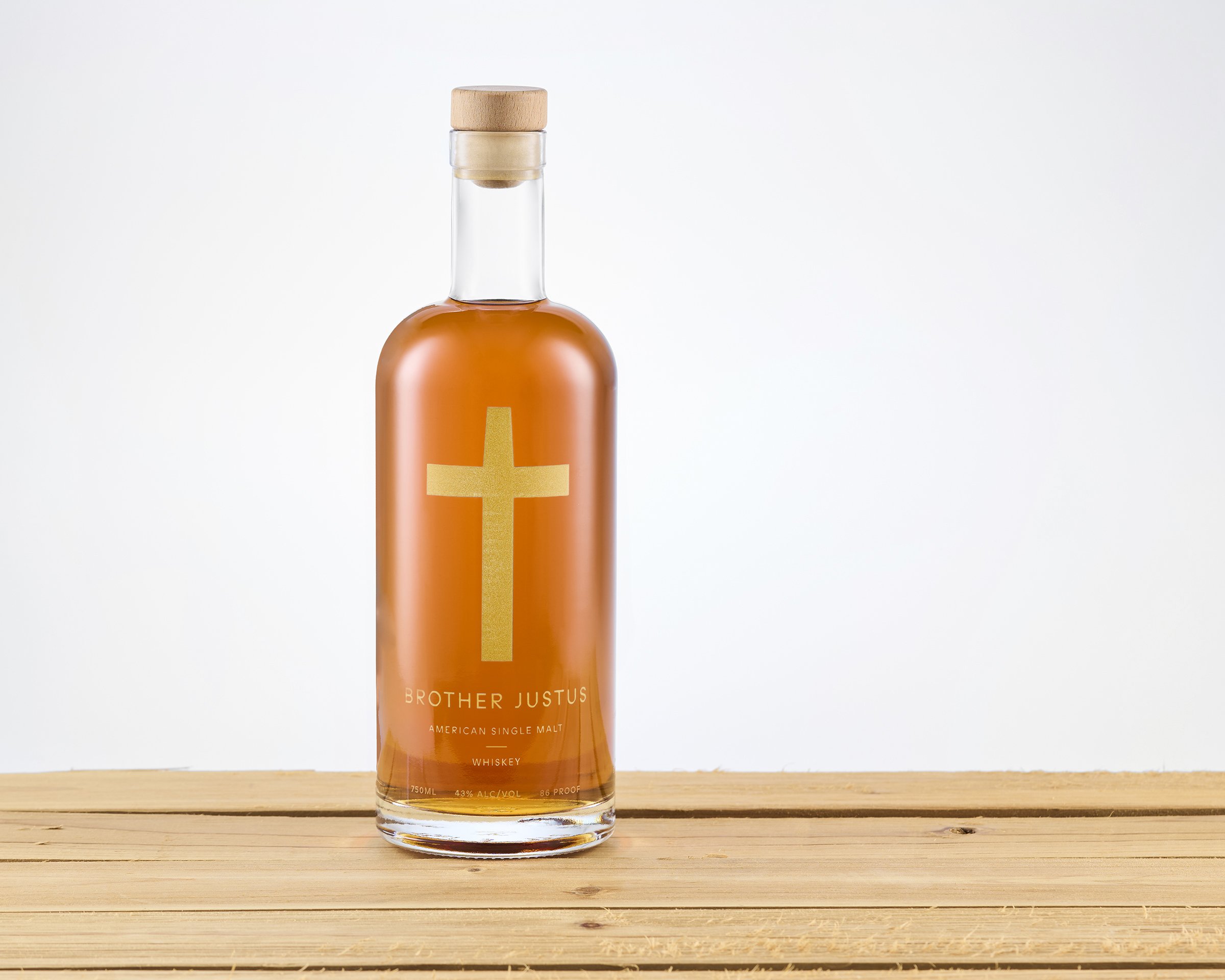 A clear glass bottle of whiskey with a wooden cap, featuring a gold cross symbol and label reading Brother Justus American Single Malt Whiskey, placed on a wooden surface against a white background.