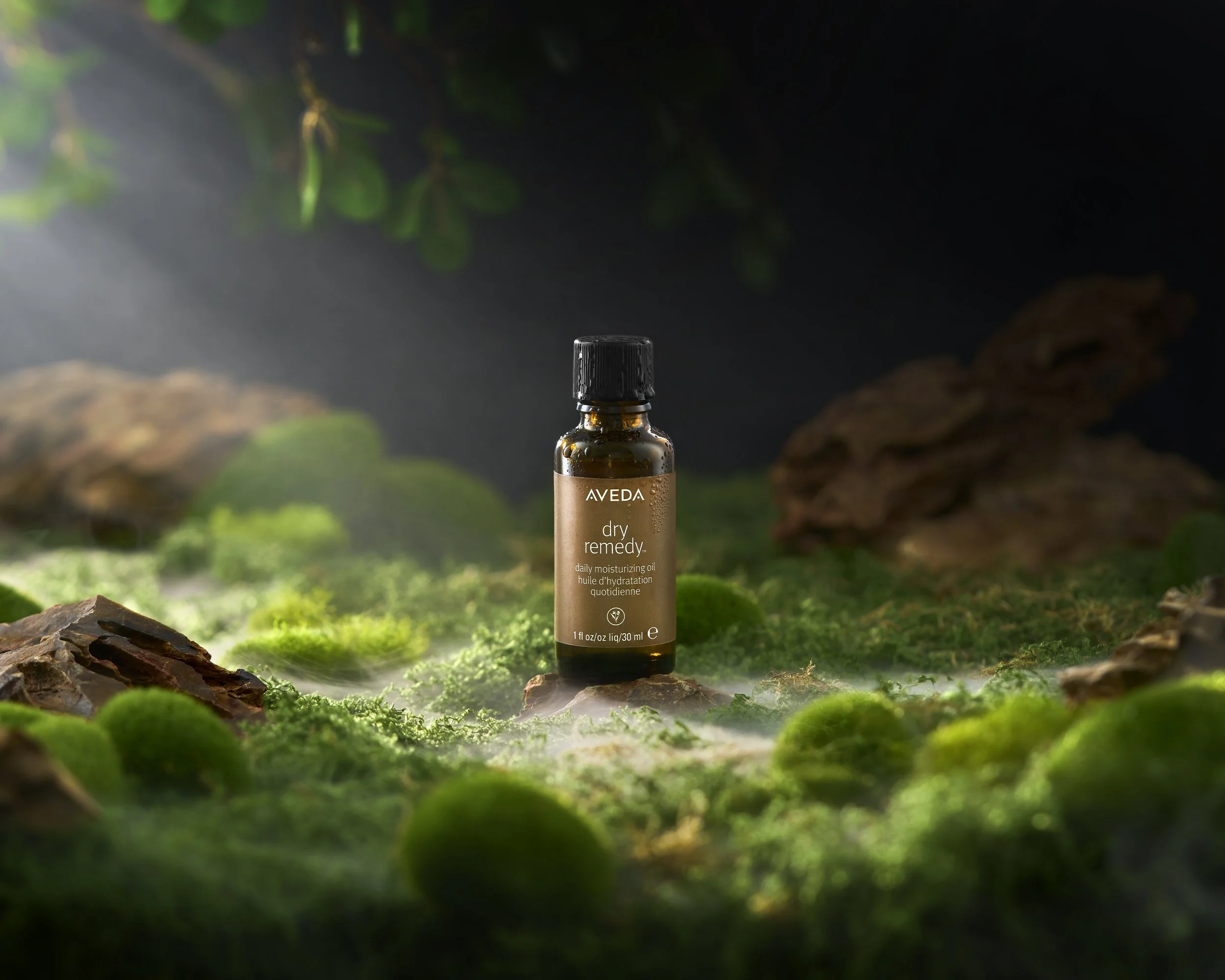 A small amber glass bottle labeled 'AVEDA dry remedy' with a black cap, standing on a moss-covered surface with rocks and greenery in the background, illuminated by soft light.