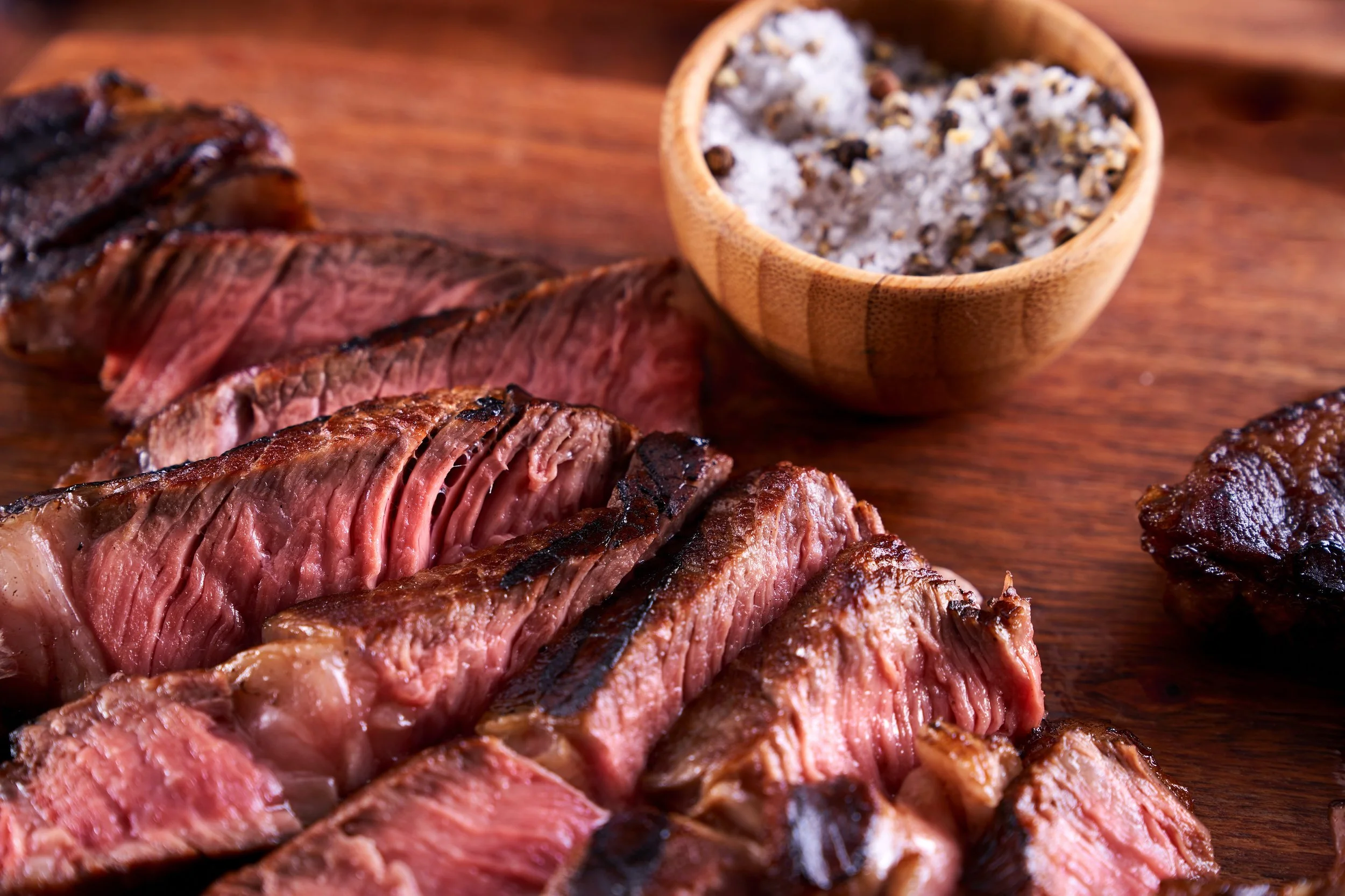 Slices of cooked steak on a wooden cutting board with a small bowl of seasoning salt.
