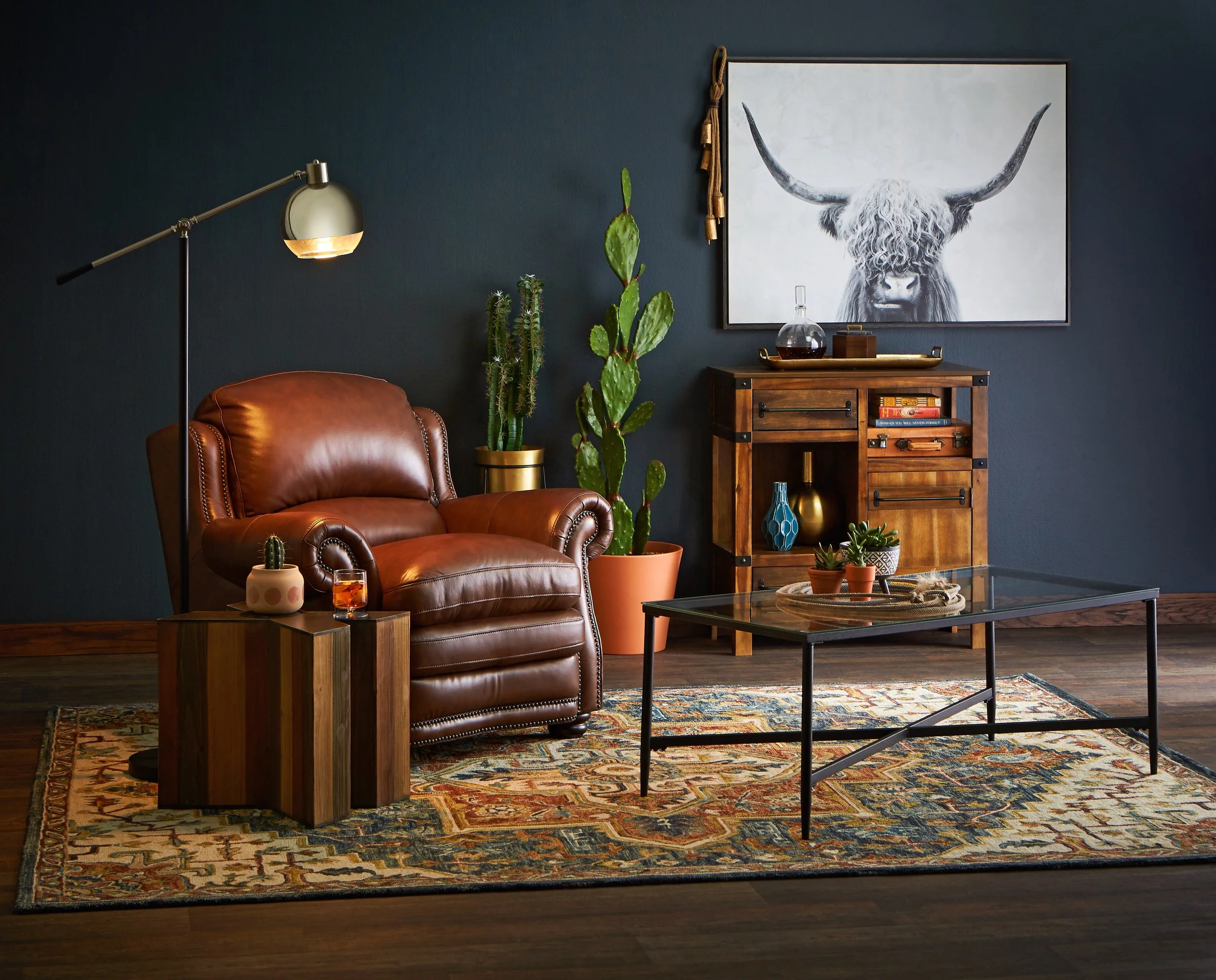 Living room with brown leather armchair, side table with cactus plant and drink, wooden cabinet with decor and books, large cactus plant, picture of a highland cow on dark blue wall, glass coffee table, patterned rug