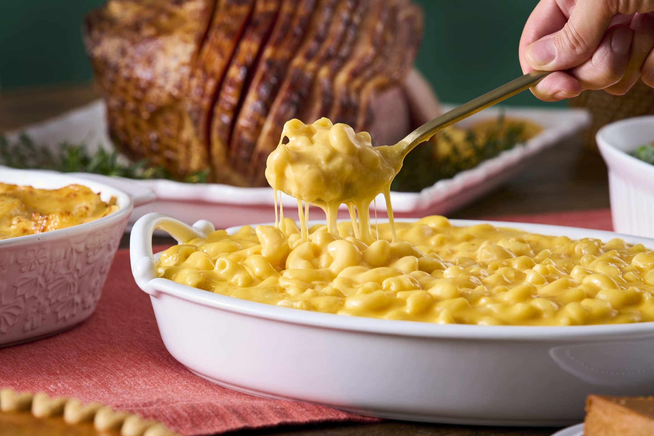 A white dish of creamy macaroni and cheese with cheese sauce dripping from a ladle, in front of a serving of glazed ham in the background.