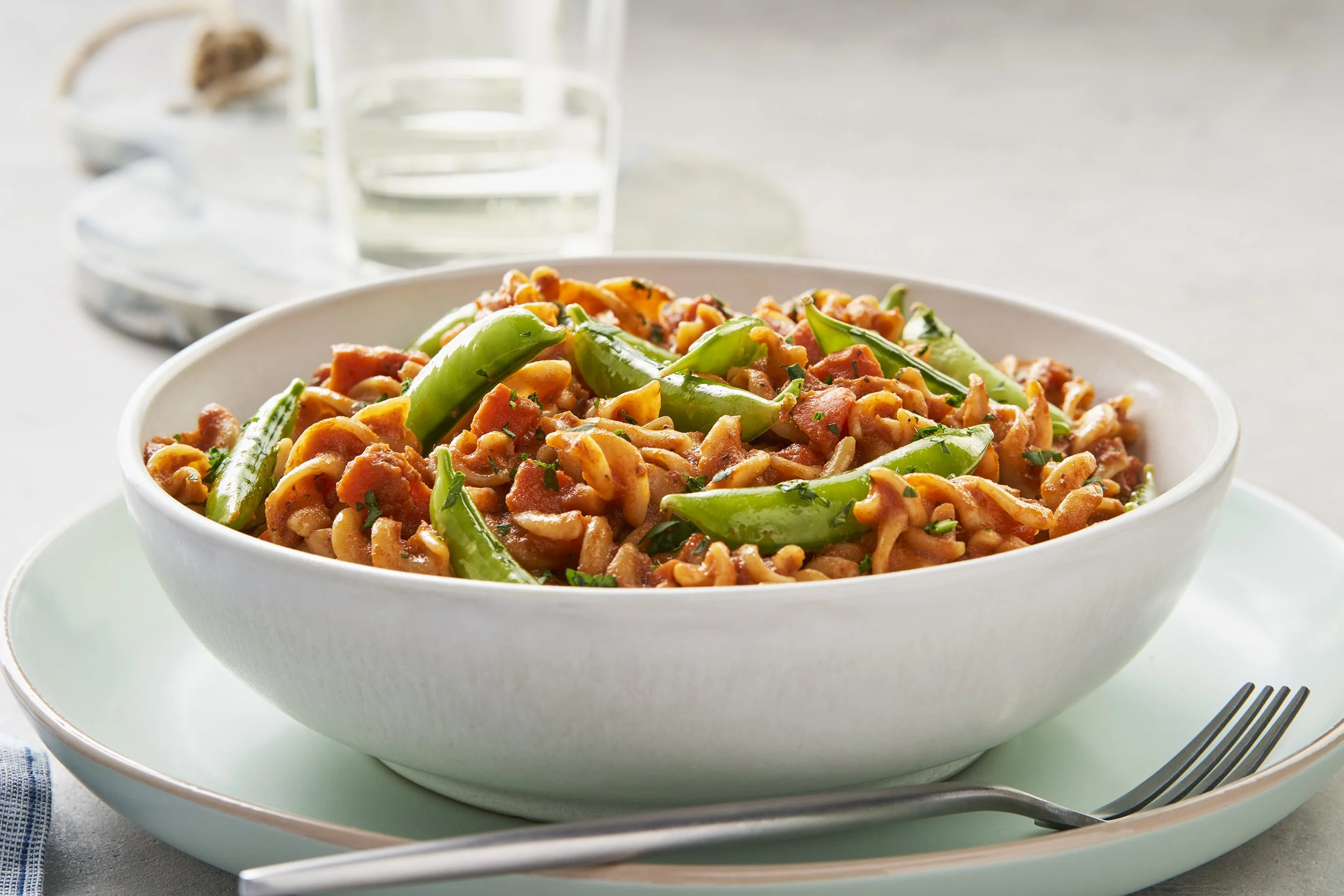 A white bowl filled with pasta and green peppers, served on a light green plate with a fork, in front of a glass of water.