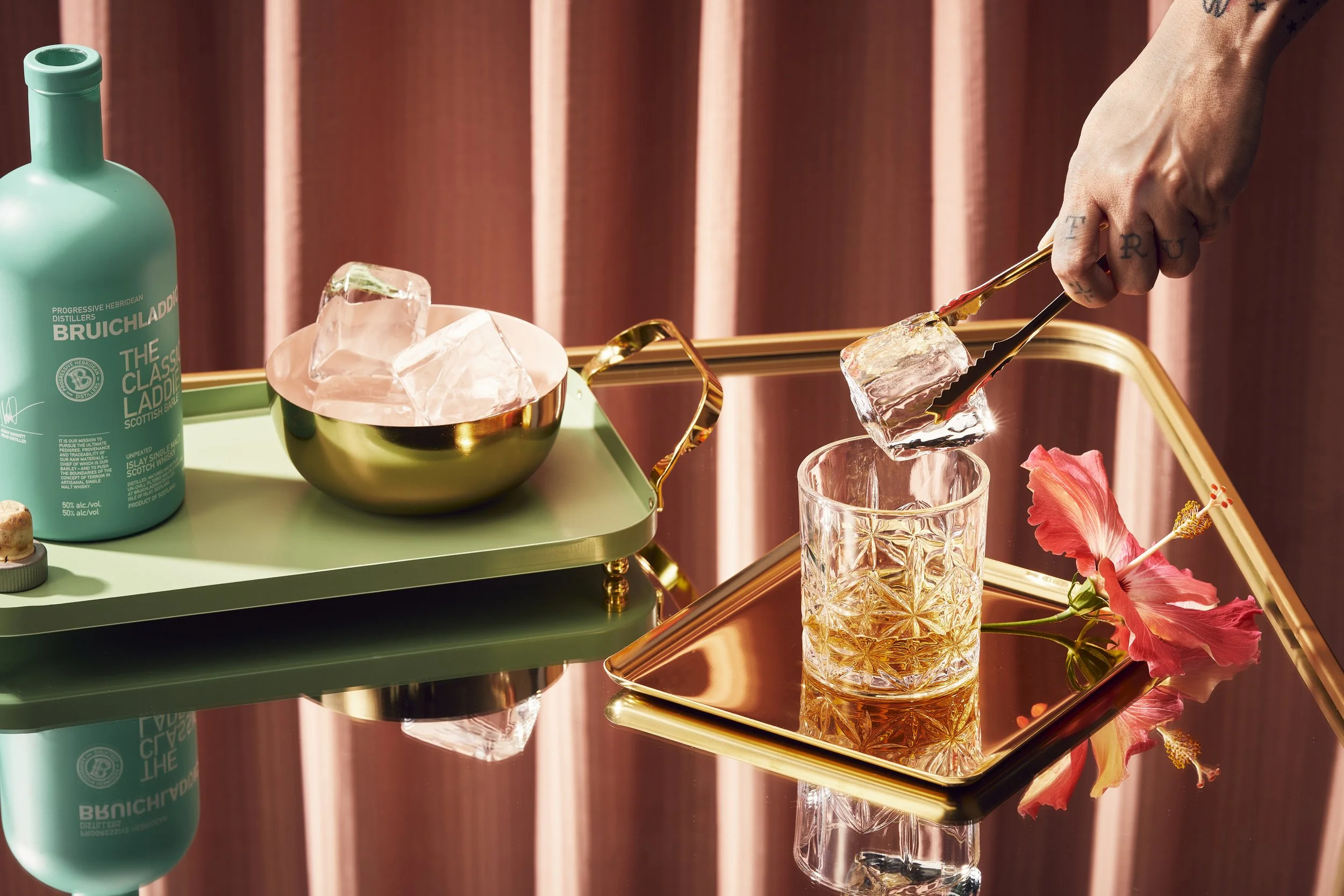 Person using tongs to place ice cubes into a glass of amber-colored drink on a reflective gold tray with pink hibiscus flower, on a table with a pink curtain background.
