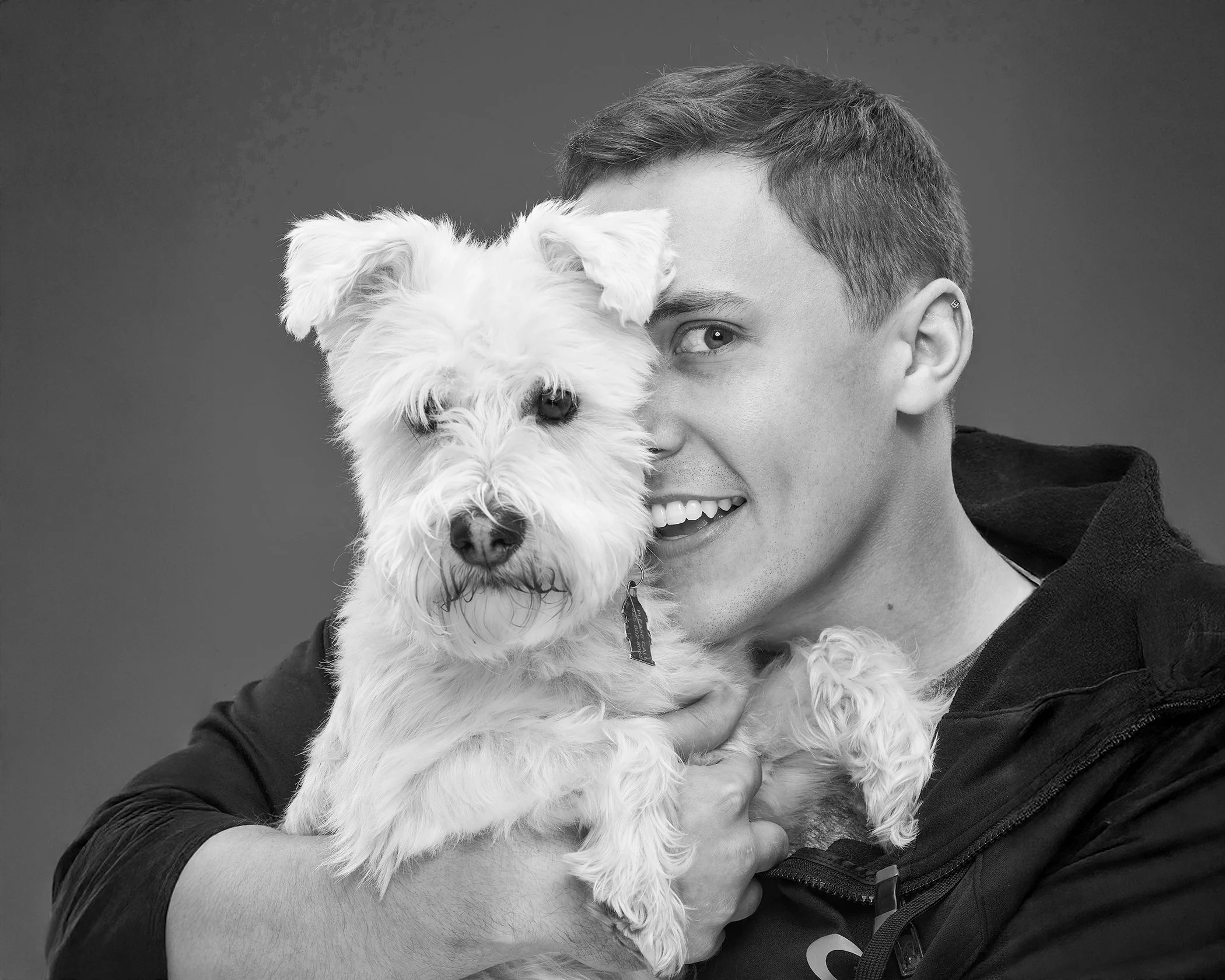 A young man holding a small fluffy dog close to his face, both looking at the camera, in black and white.