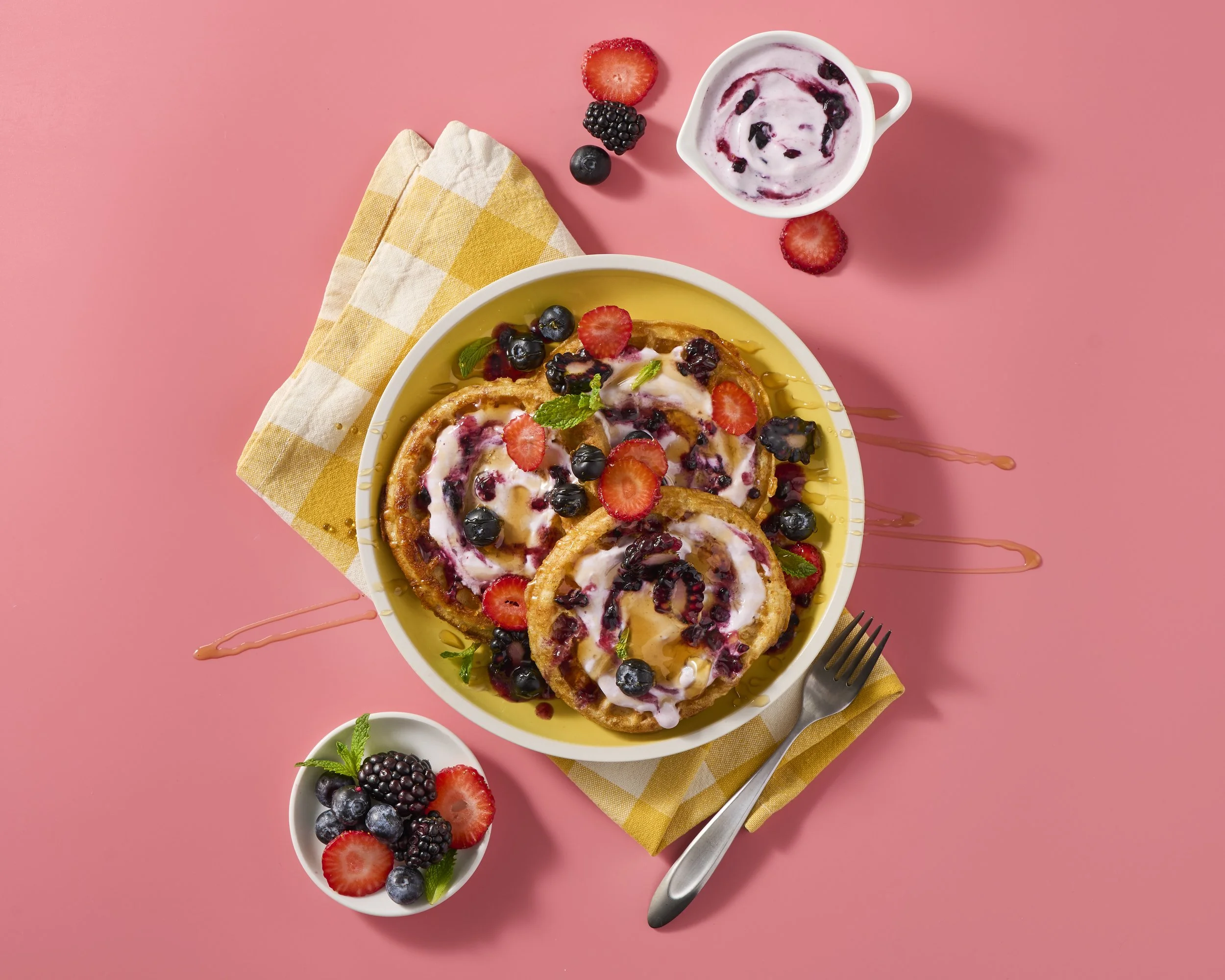 French toast topped with mixed berries, whipped cream, and mint leaves, served with yogurt and fresh berries on the side, on a pink background.