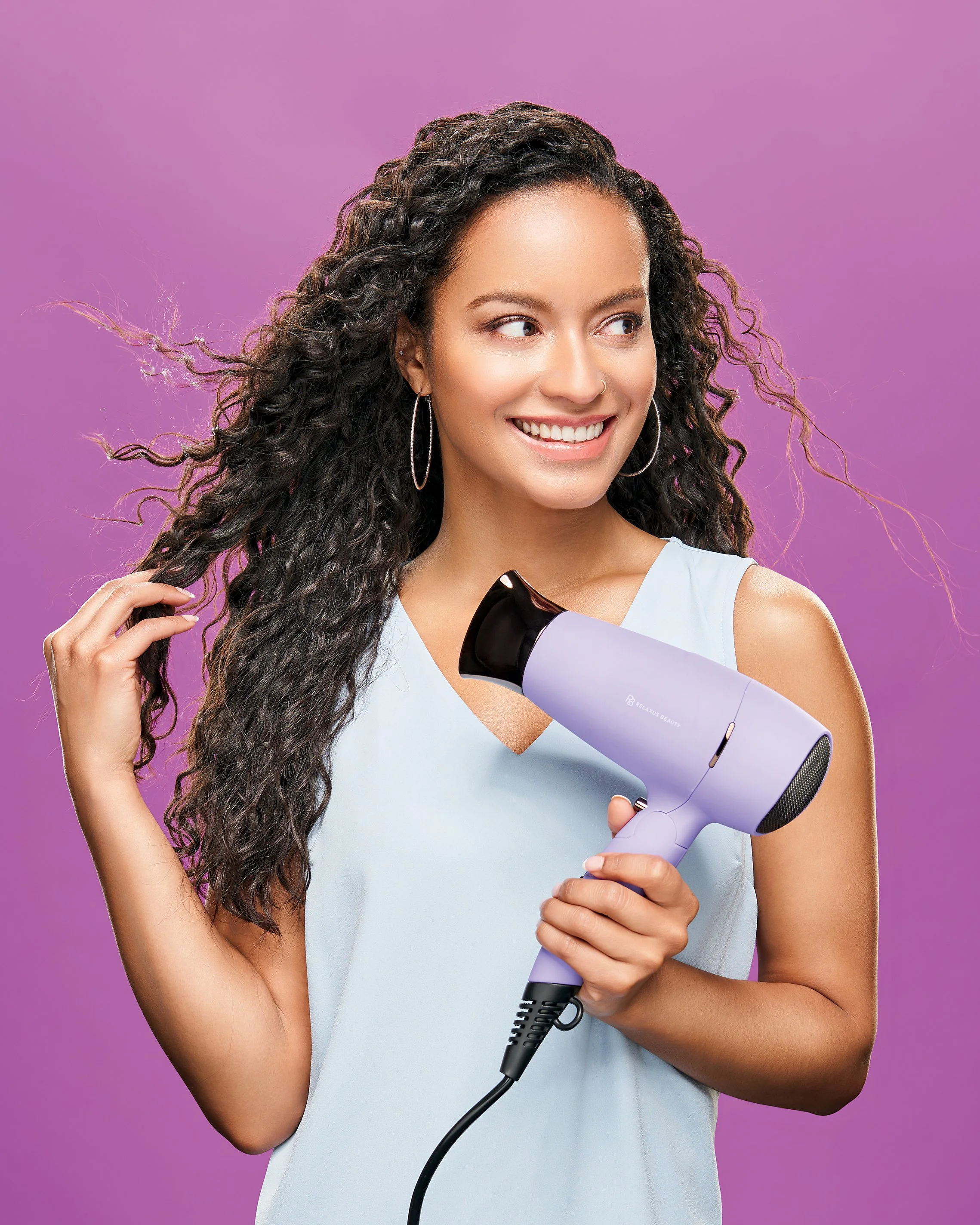 A woman with curly hair styling her hair with a purple hair dryer against a purple background.