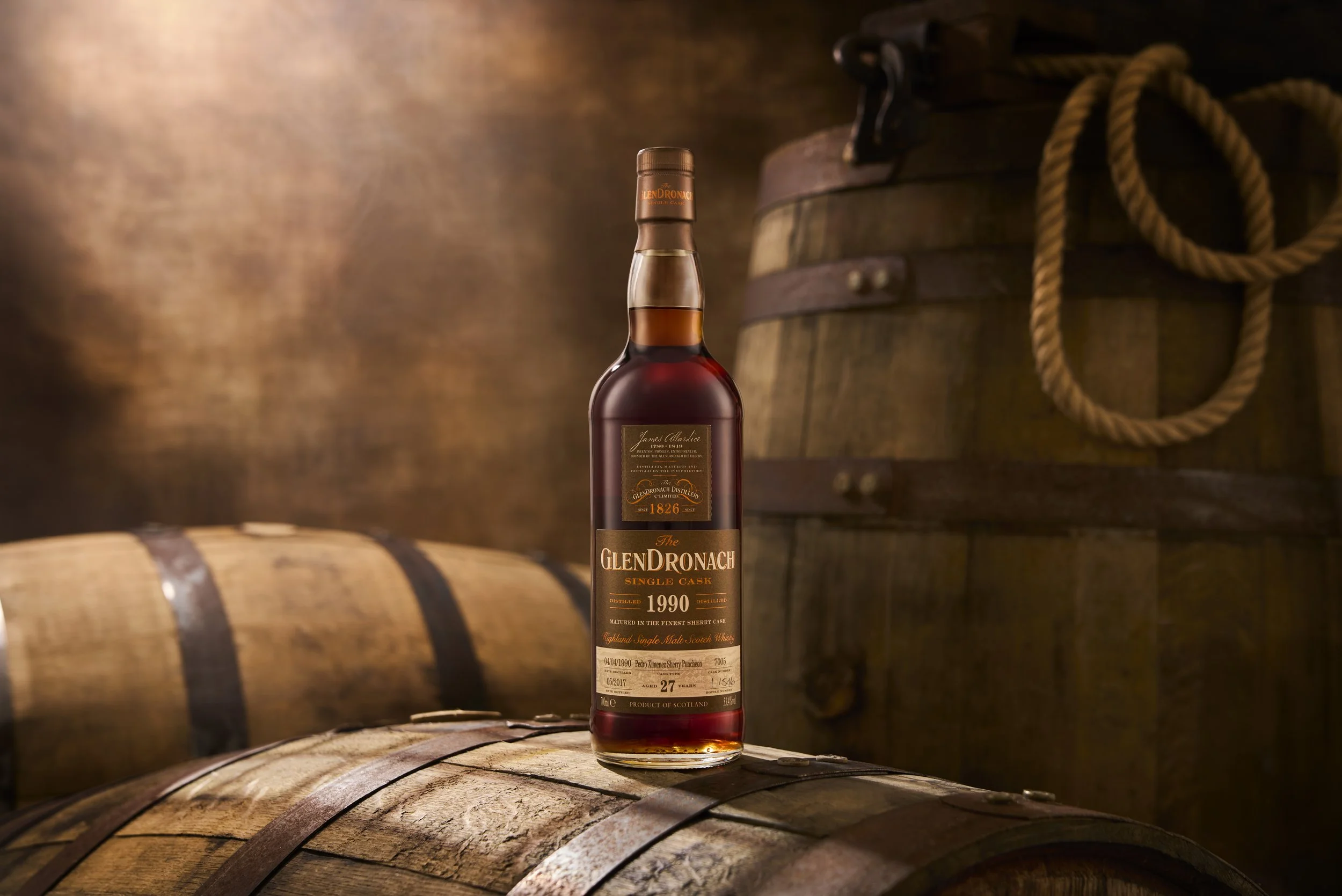 A bottle of Glendronach single malt Scotch whisky from 1990 is placed on a wooden barrel in a dimly lit space with another barrel and coiled rope in the background.
