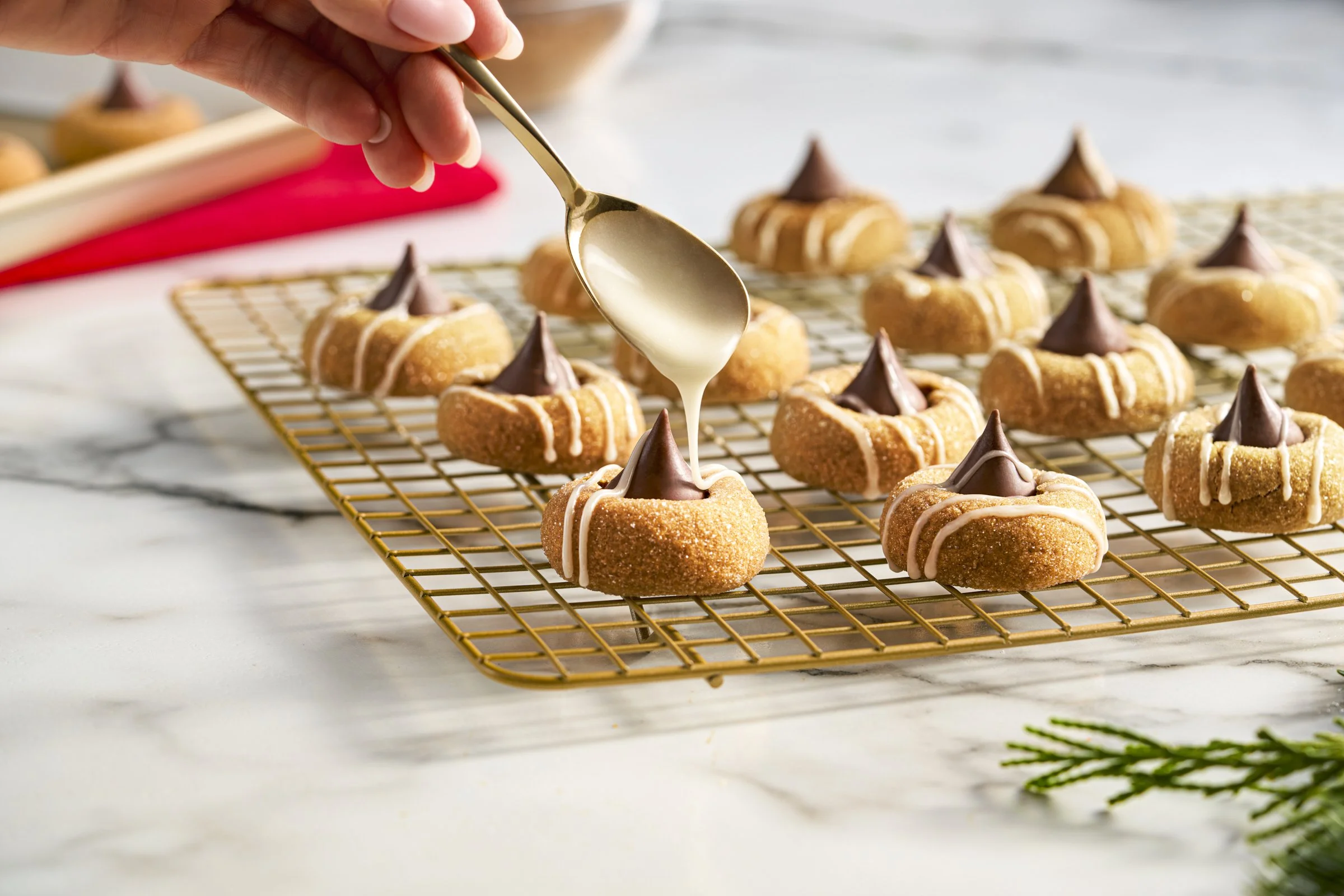Hand pouring white icing or glaze onto mini baked goods with chocolate topping, placed on a cooling rack on a marble countertop.