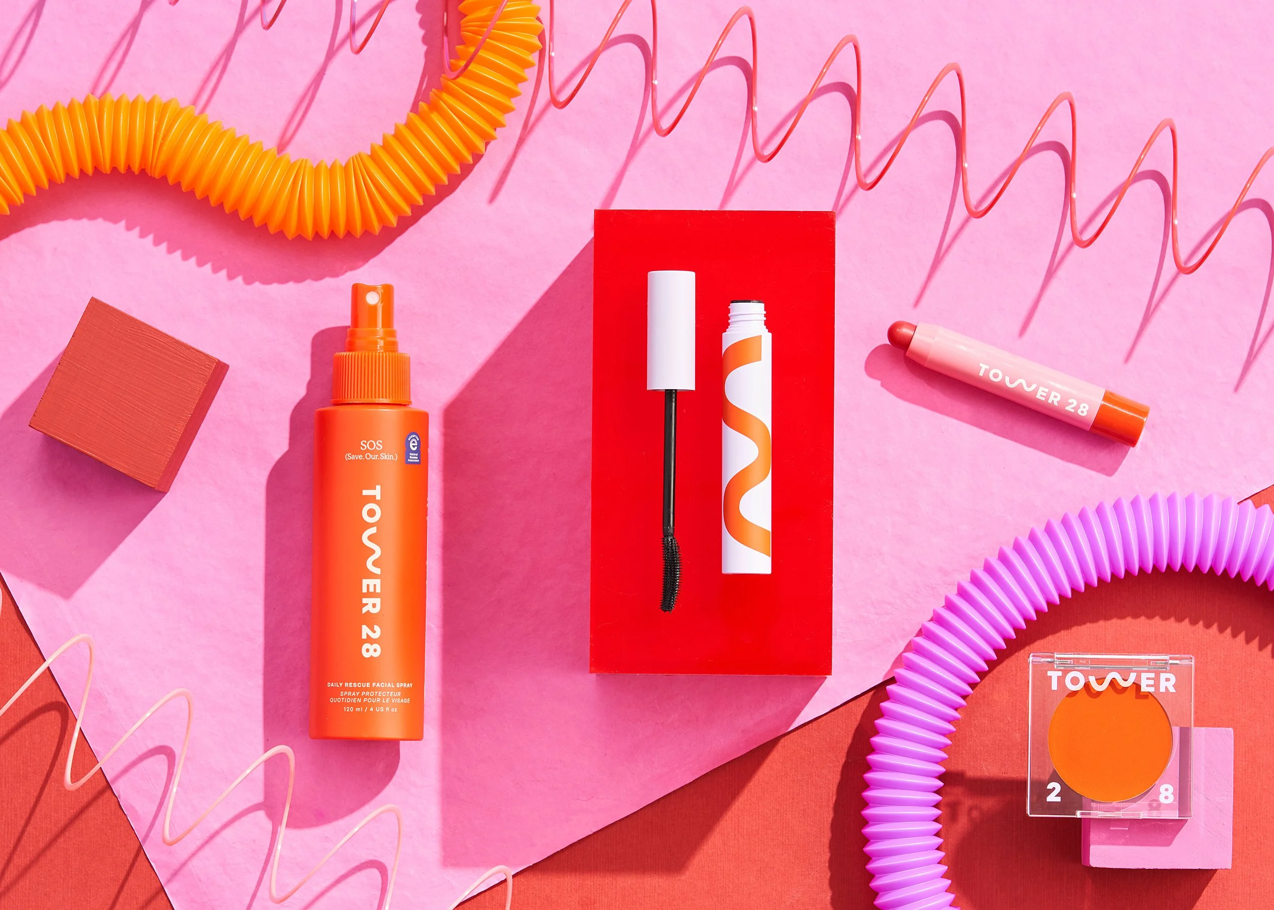 Various beauty and skincare products in bright orange and pink packaging, arranged on a pink background with colorful decorative elements including a spiral tube, a spray bottle, a mascara wand, and a small square container.