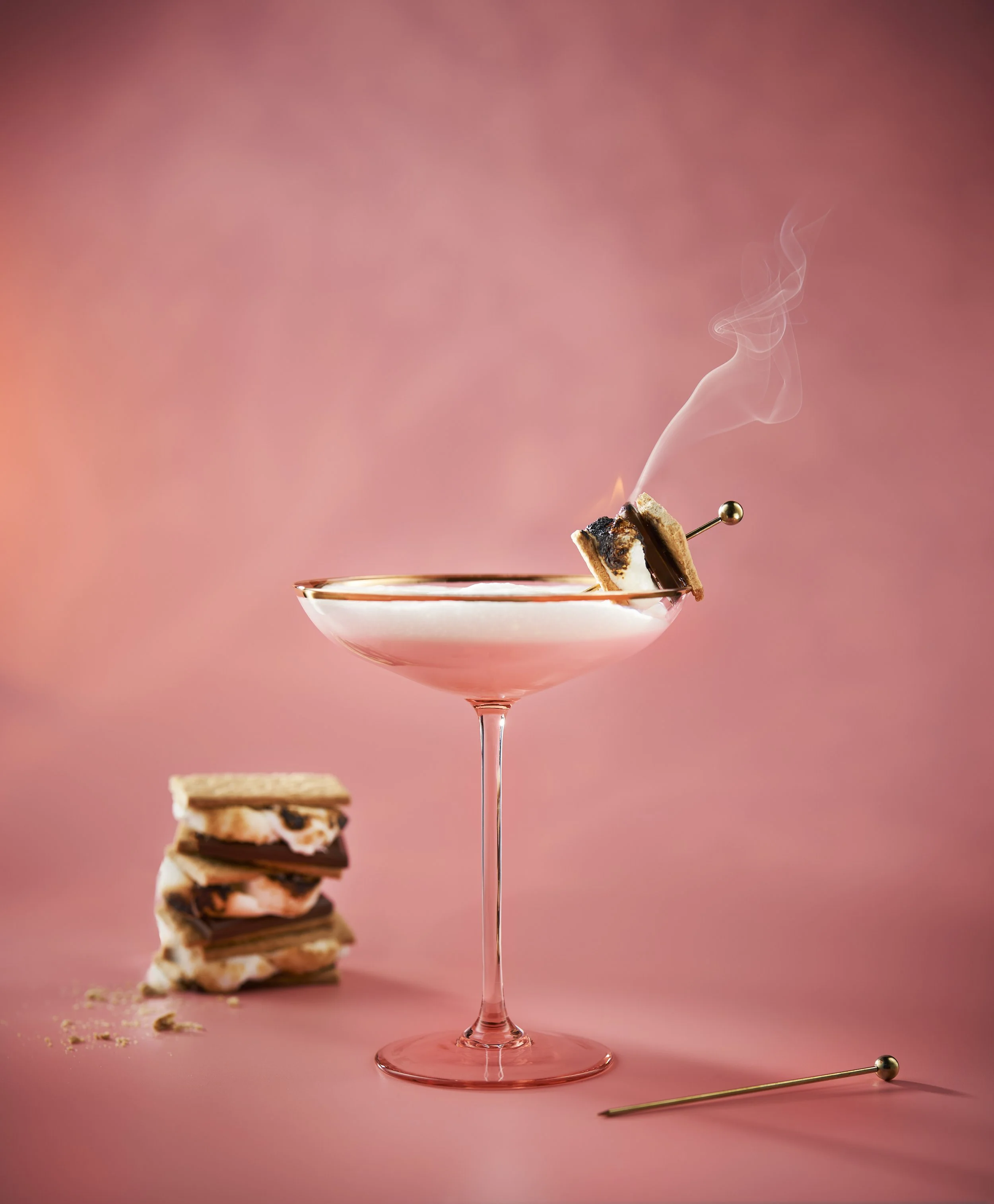 A pink cocktail glass with a smoked dessert on top, a small stack of s'mores on the side, and a pin on the drink's rim, set against a pink background.