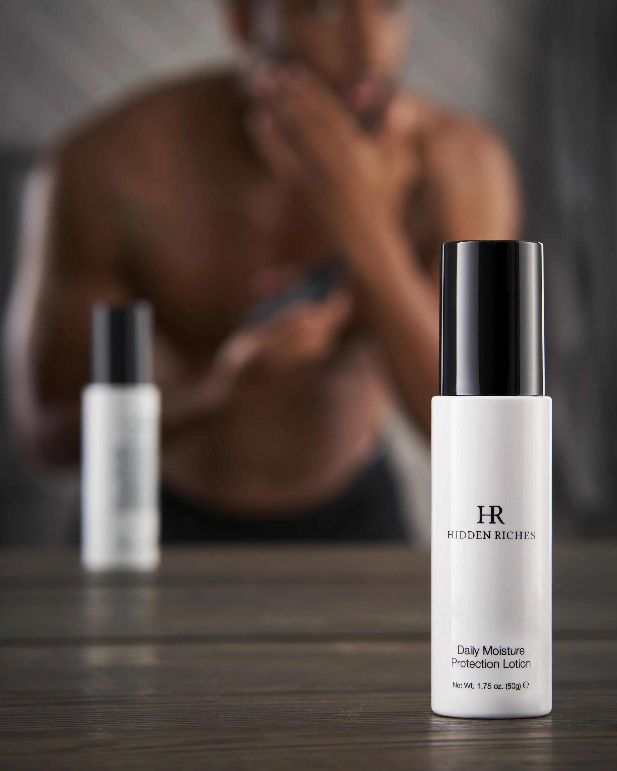 A bottle of Hidden Riches Daily Moisture Protection Lotion on a wooden surface with a blurred woman in the background applying lotion to her shoulder.