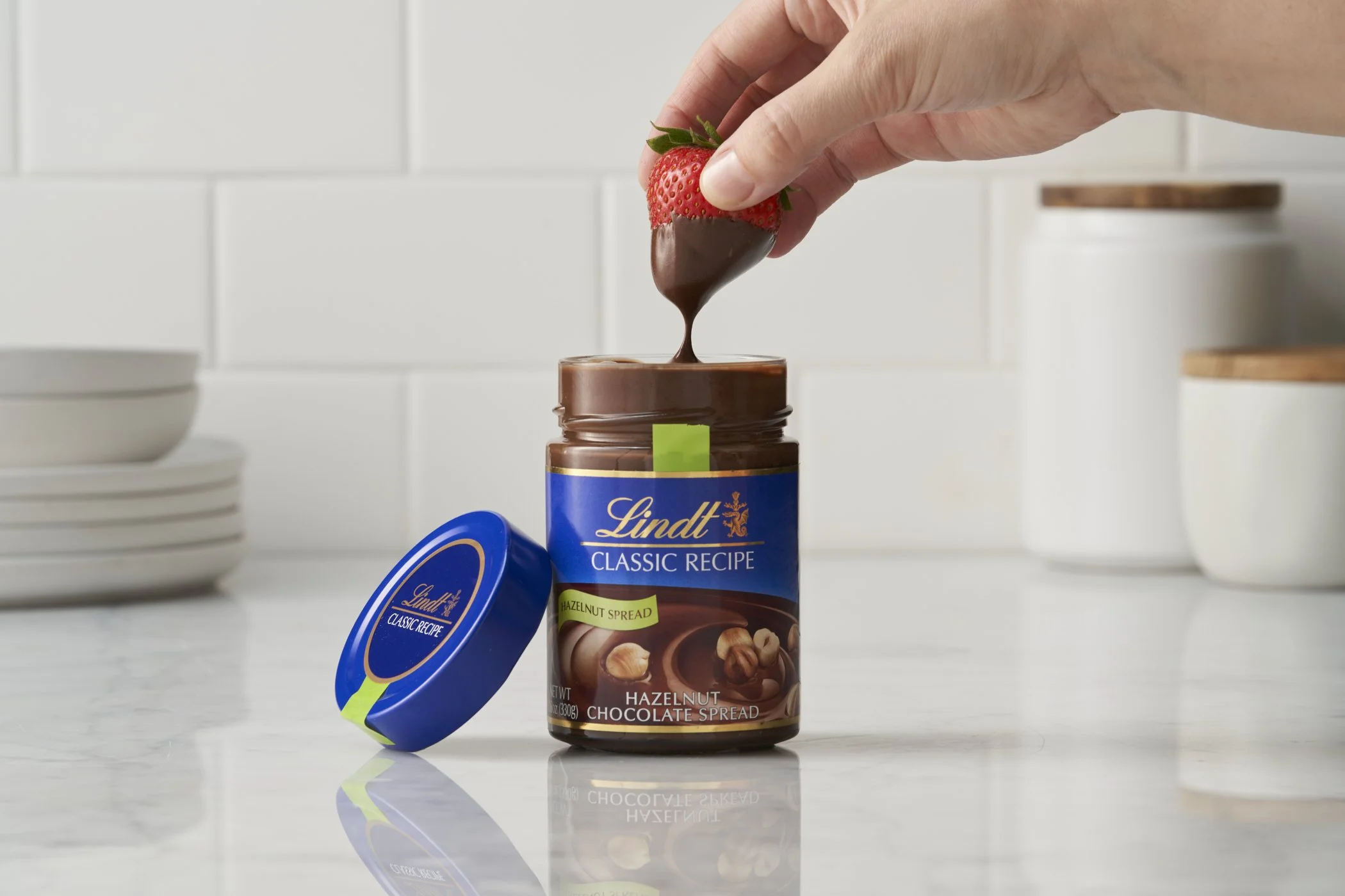 A hand dipping a fresh strawberry into chocolate hazelnut spread from a jar of Lindt Classic Recipe Hazelnut Chocolate Spread with the jar's lid placed on the countertop.