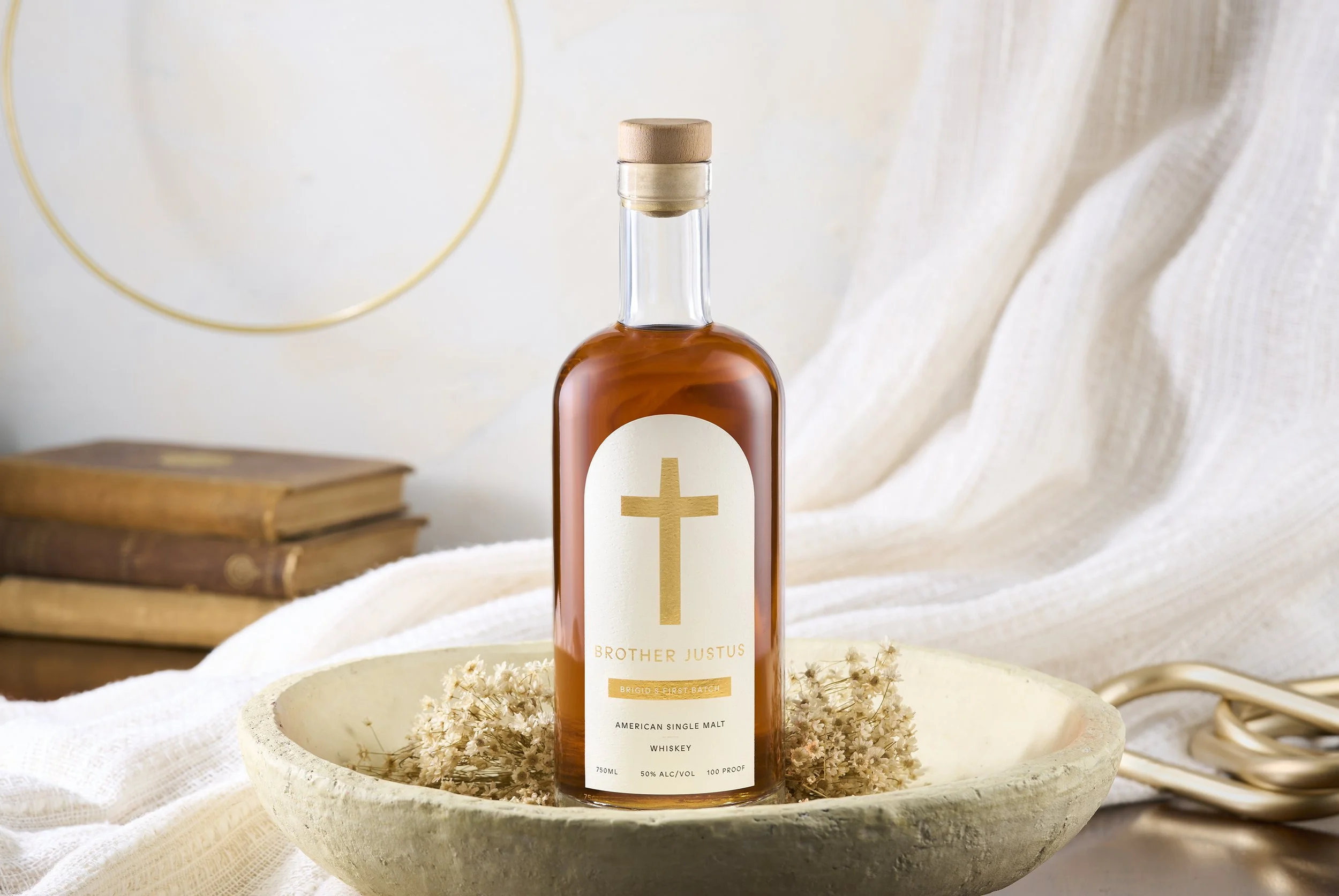 A bottle of whiskey with a label featuring a gold cross and the words 'Brother Justus' is placed on a shallow bowl filled with dried flowers, with a stack of old books, a white cloth, and a pair of scissors in the background.